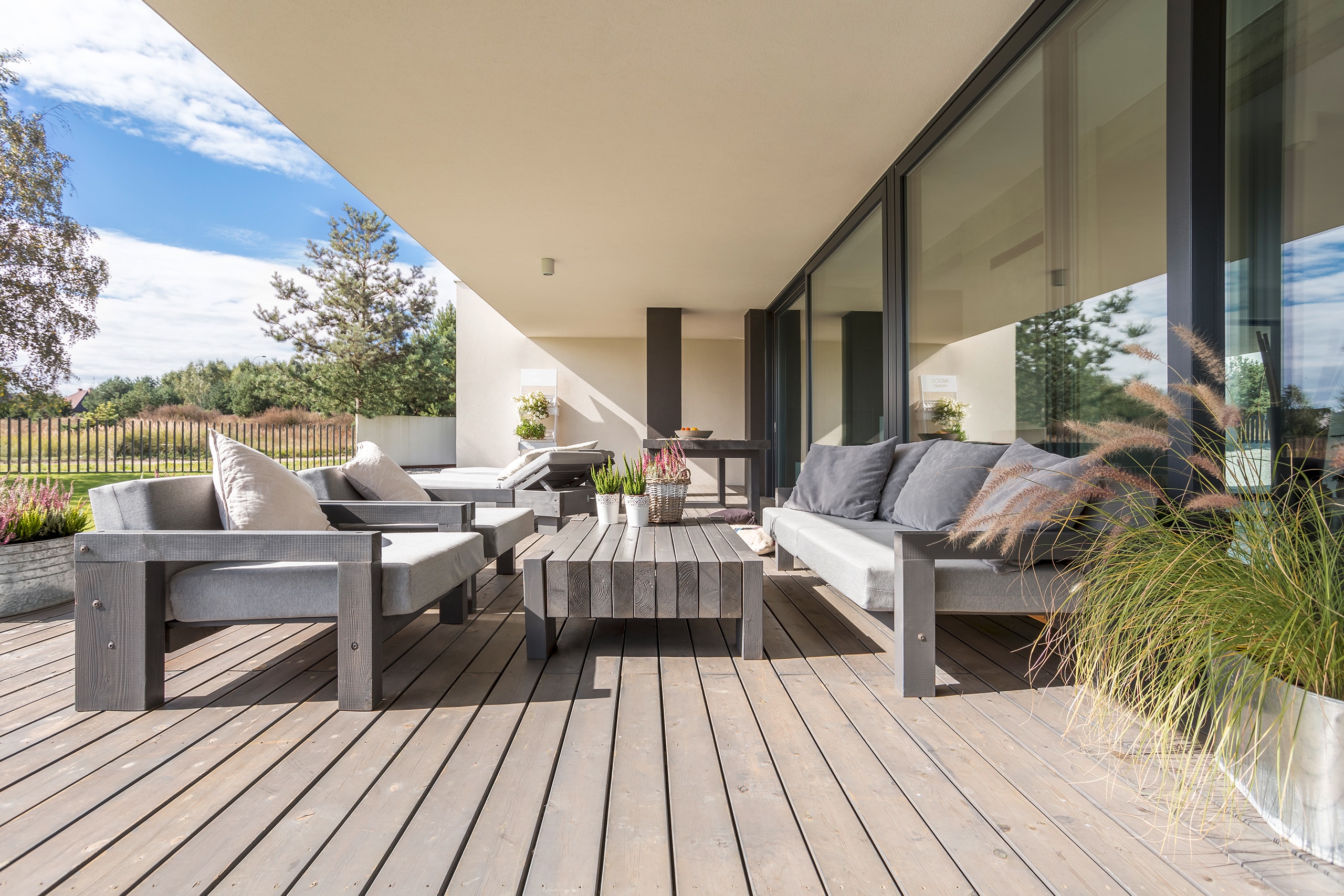 Terrassenbereich mit Holzdielen, darauf moderne, gemütliche Lounge-Gartenmöbeln in Grau, witterungsgeschützt unter dem Vorsprung eines modernen Hauses mit großer Fensterfront.