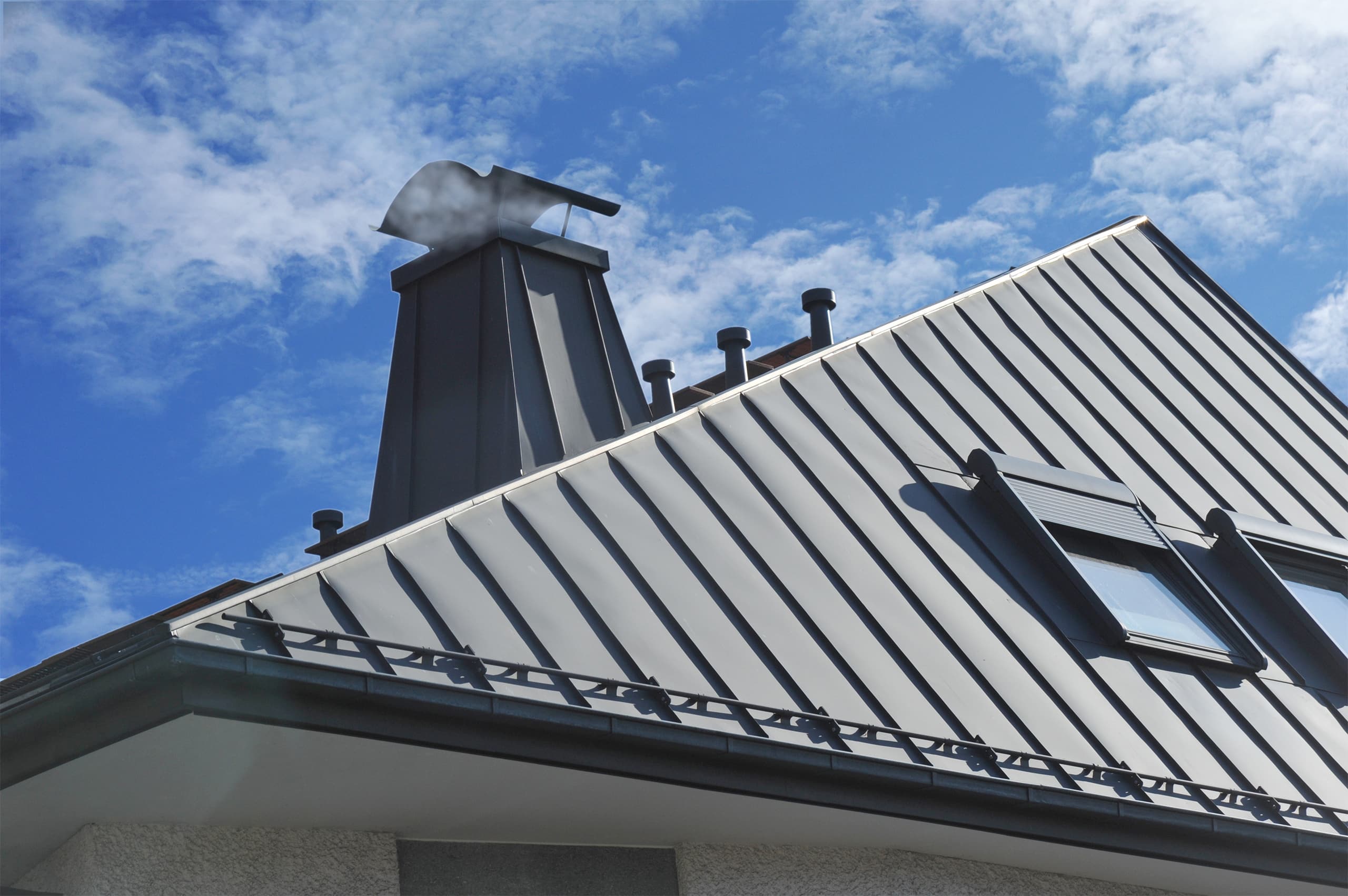 Modernes Stehfalz-Metalldach mit Dachfenster, Schneefang, Dachrinne, Dunstabzügen und mit Metall verkleidetem Schornstein (Rauchfang) am Dach mit einer dunklen Blecheindeckung.