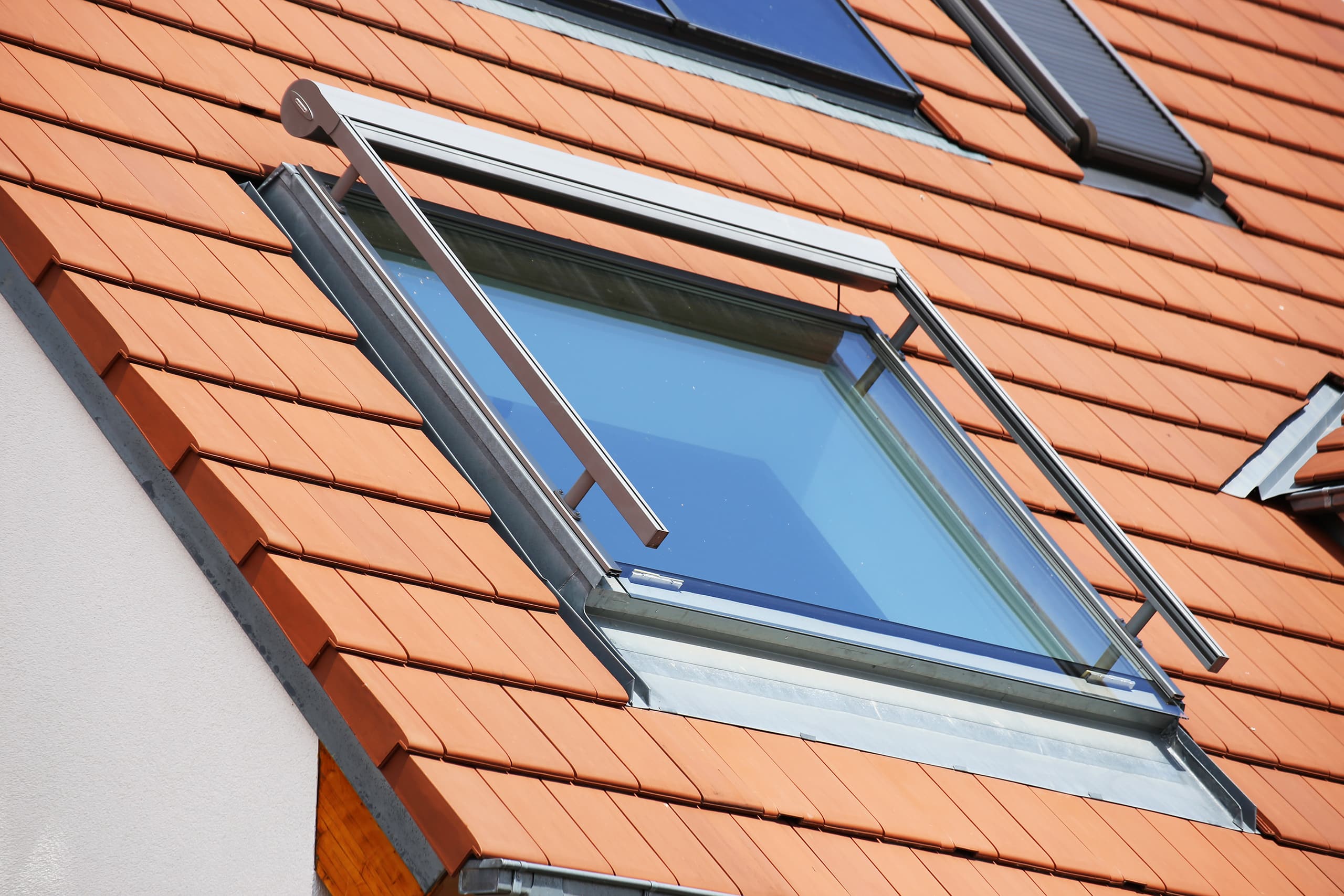 Detailfoto eines Daches mit rotern Dachziegeln sowie quadratischem Dachflächenfenster mit Markise.