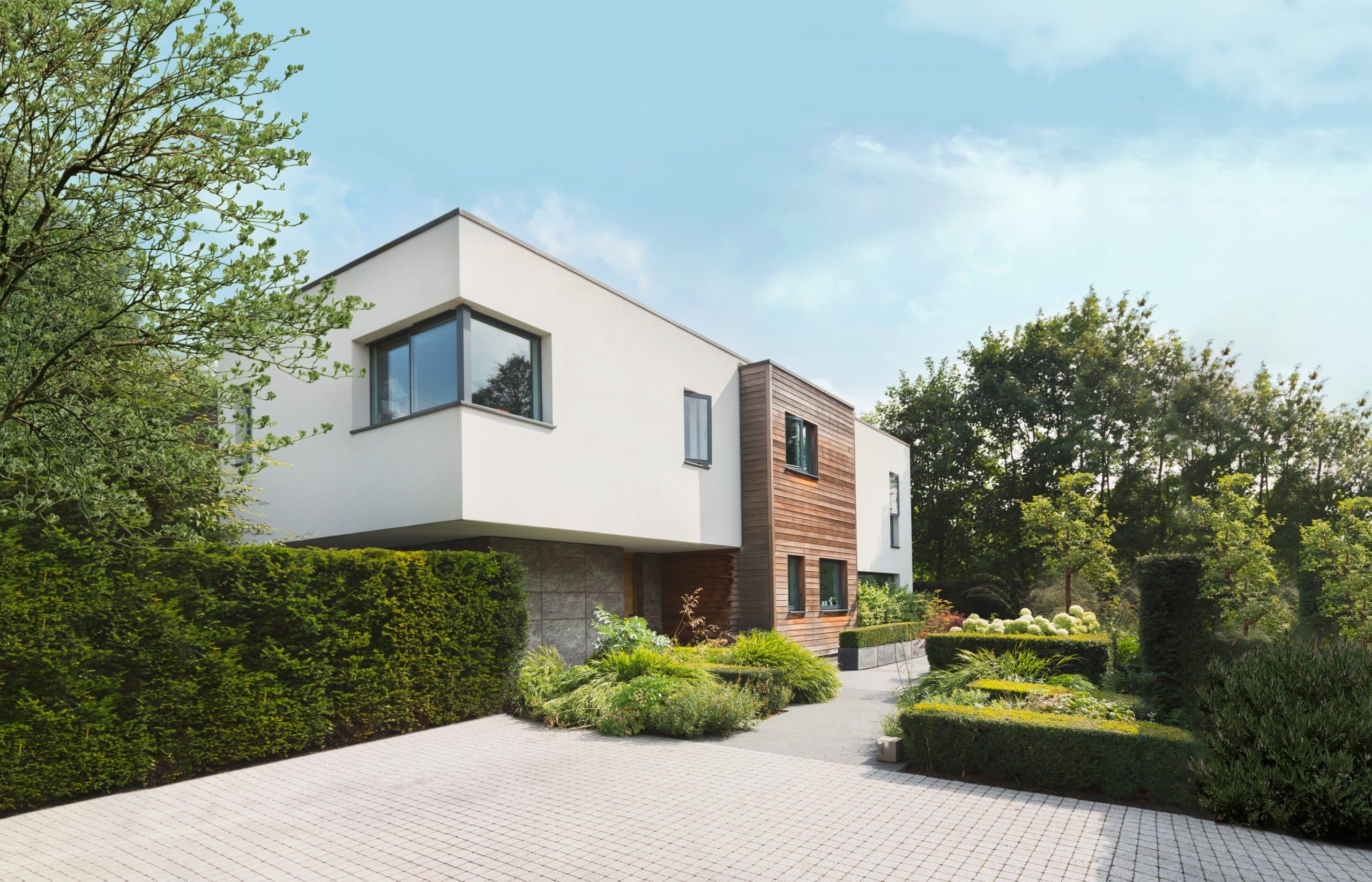 Modernes Haus mit weißer Fassade, unterbrochen durch Holzelemente mit ansprechender Gartengestaltung inklusive Bäumen, Sträuchern und Hecken sowie weiß blühenden Hortensien.
