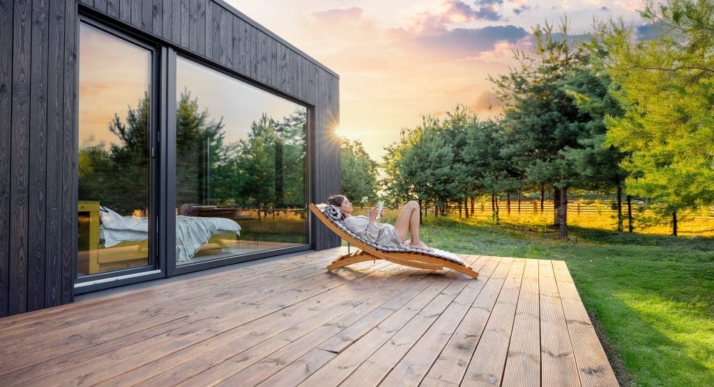 Junge Frau liegt gemütlich im hölzernen Liegestuhl auf der Terrasse mit Holzdielen und dem modernen Haus mit Holzfassade und Ganzglas-Schiebetüren und genießt die Abendsonne mit dem Lesen eines Buches.