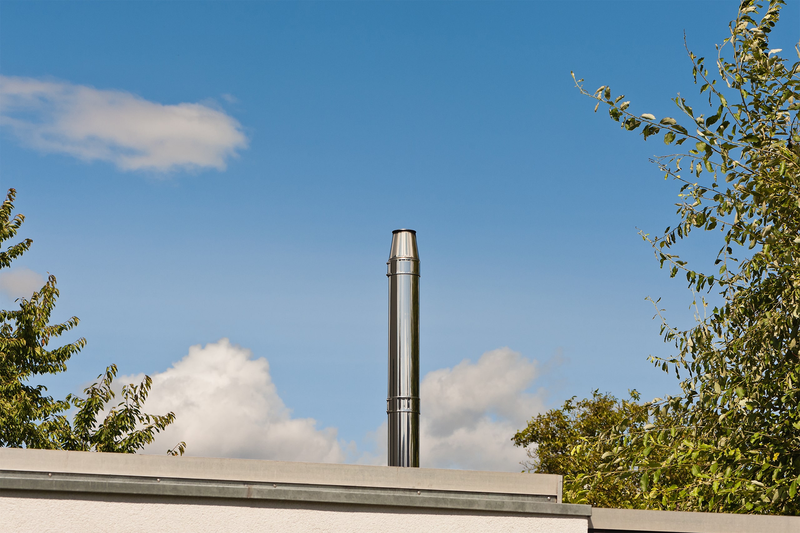 Schornstein (Rauchfang) aus Edelstahl auf einem Flachdach, der in den sonnigen Himmel ragt.
