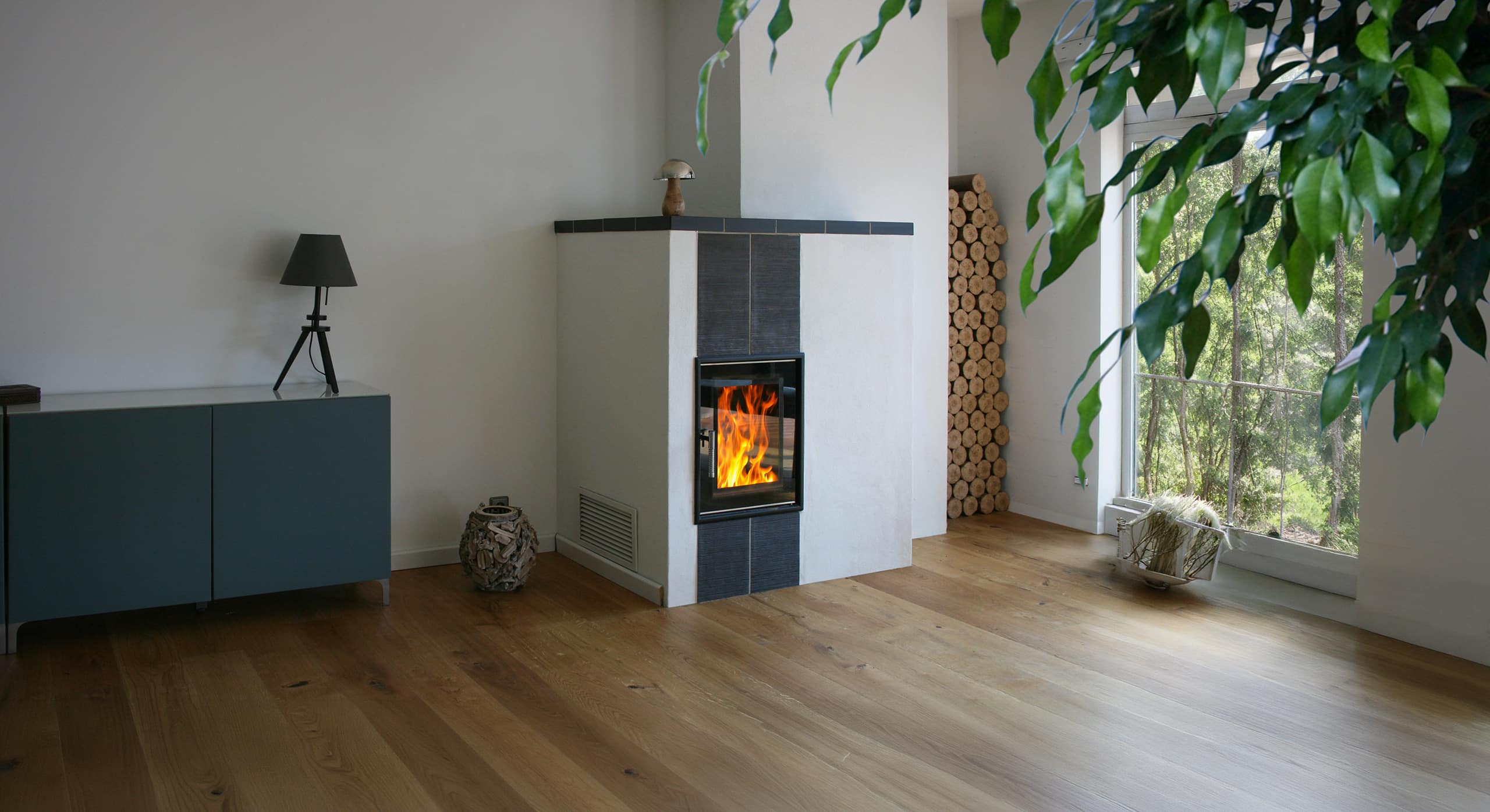 Moderner Kachelofen mit loderndem Feuer durch die Glasscheibe und Holzbriketts zum Nachheizen in einer Wandniesche neben großem Fenster mit herrlichem Blick in die grüne Natur.