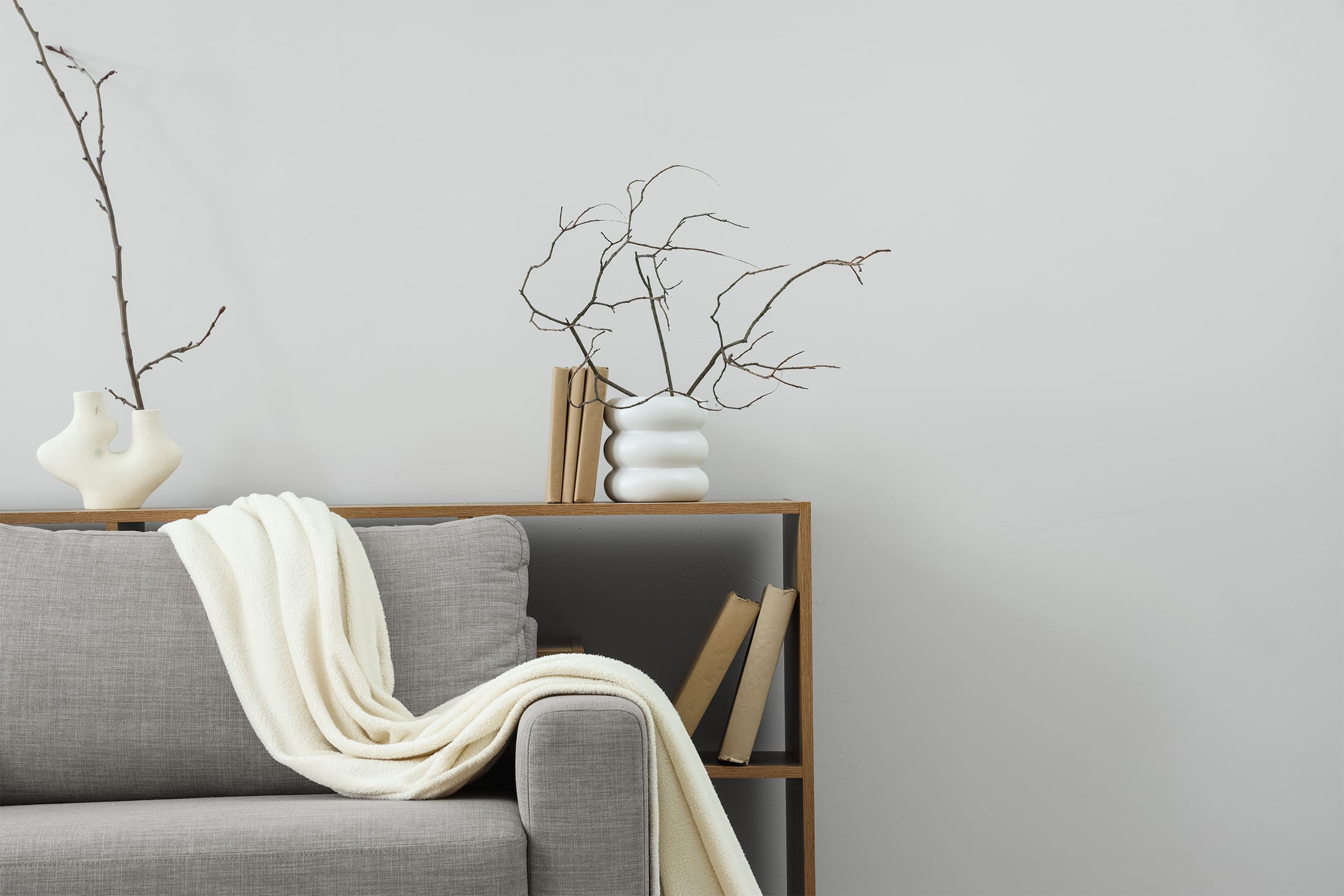Bücher und Vasen mit getrockneten Zweigen in einem Regal aus hellem Holz im Wohnzimmer. Davor steht ein bequemes Sofa in Grau, mit einer weißen Tagesdecke trappiert.
