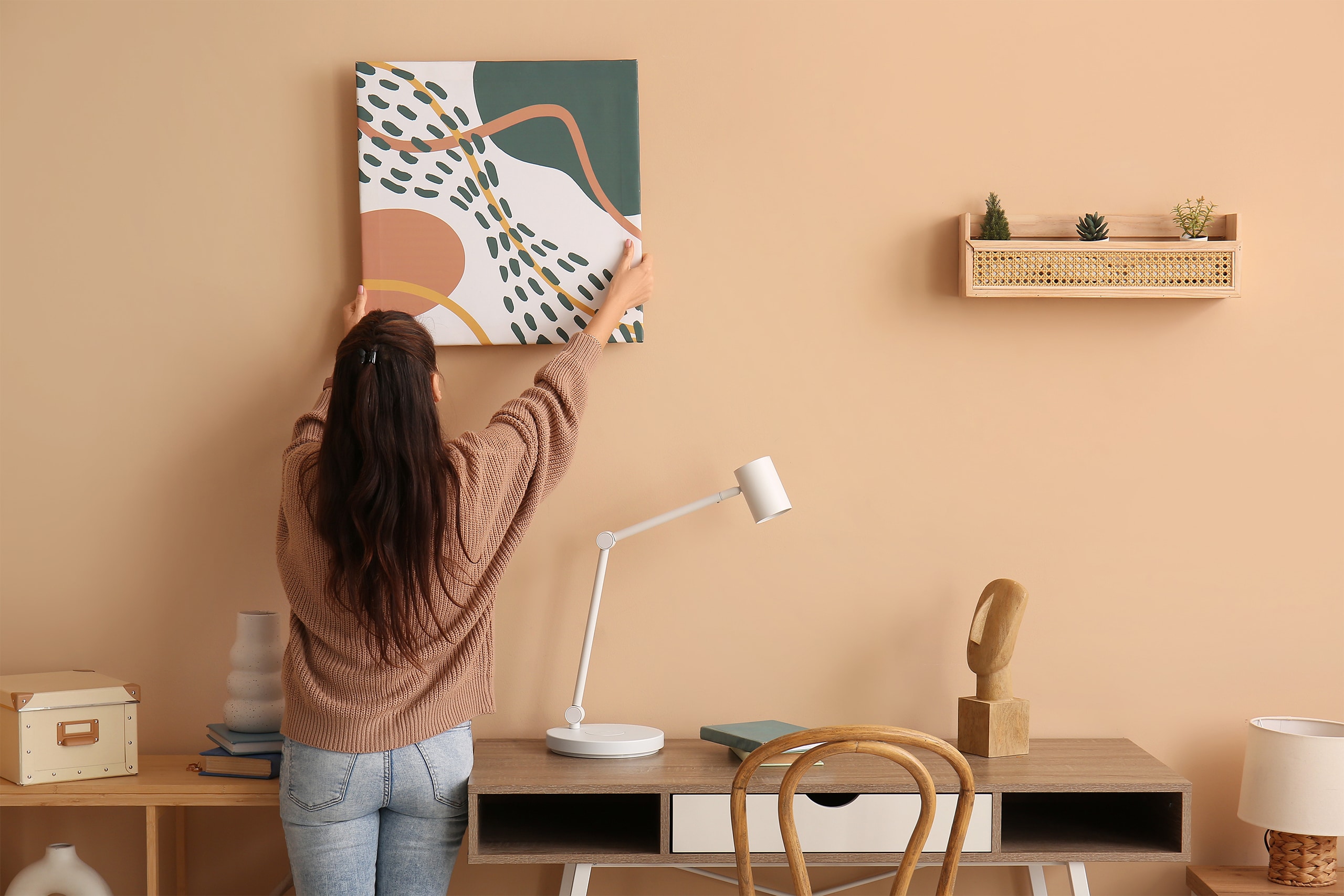 Junge Frau hängt ein selbst gemaltes Bild an die bunte, fröhliche Wand Ihrer Wohnung. Darunter befindet sich ein Schreibtisch aus Holz, darauf eine weiße Stehlampe und Accessoires.