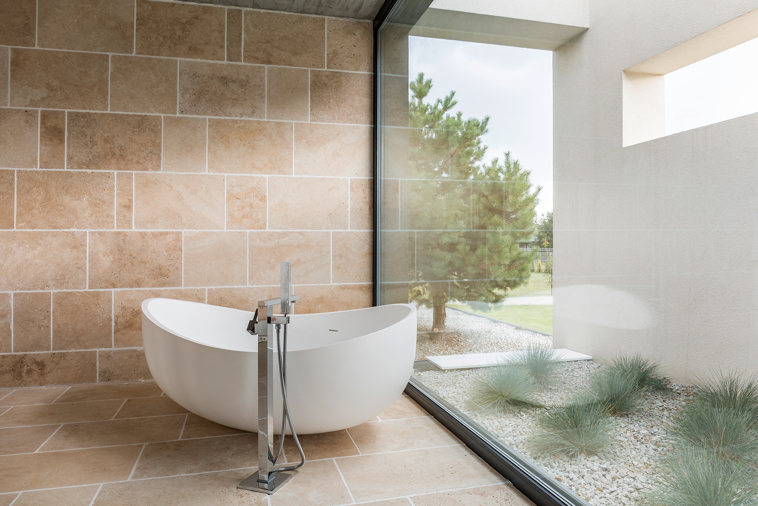 Freihstehende, weiße Badewanne mit moderner Bodenarmatur in Bad mit beigefarbenen Boden- und Wandfliesen vor großer Glasfläche mit Blick in den Garten.
