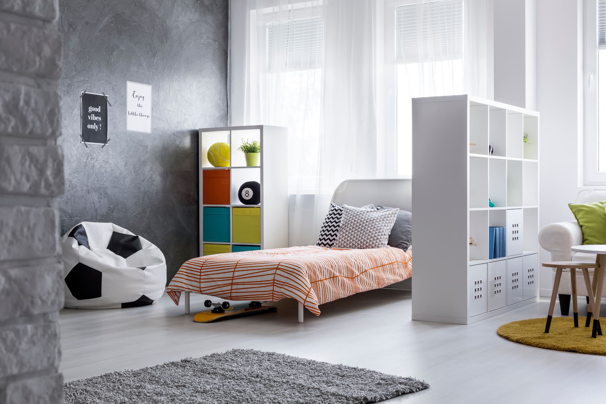 Modernes Jugendzimmer mit einem offenen Regal das als Trennwand zwischen Schlafbereich mit Bett, Kasten und Sitzsack und Wohnbereich mit Couch dient.