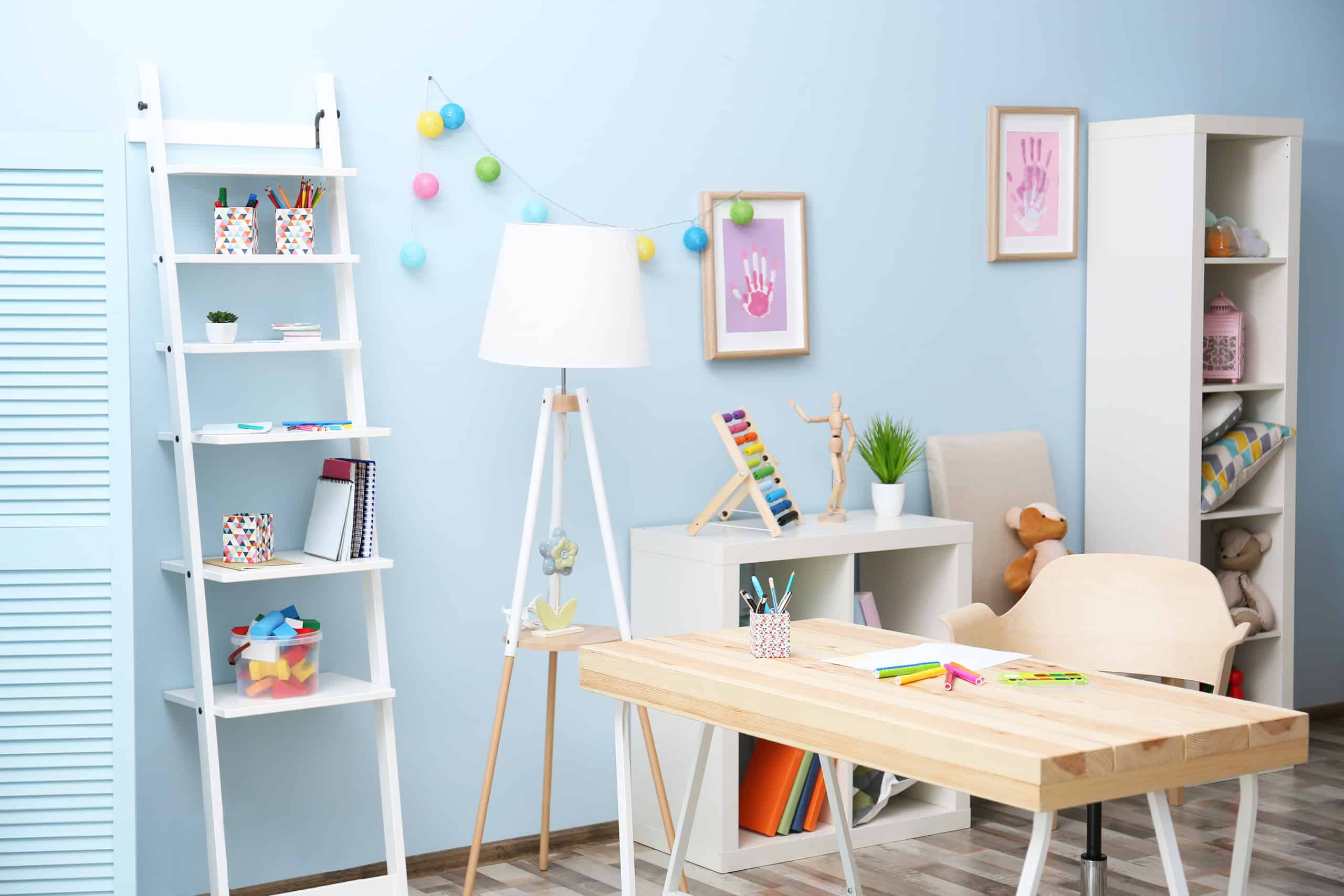 Helles Kinderzimmer mit blauer Wand, an dem ein schräges Regal lehnt, einer Stehlampe neben dem Schreibtisch, der in der Mitte des Raumes steht, einem offenen Sideboard und offenen Hochregal.