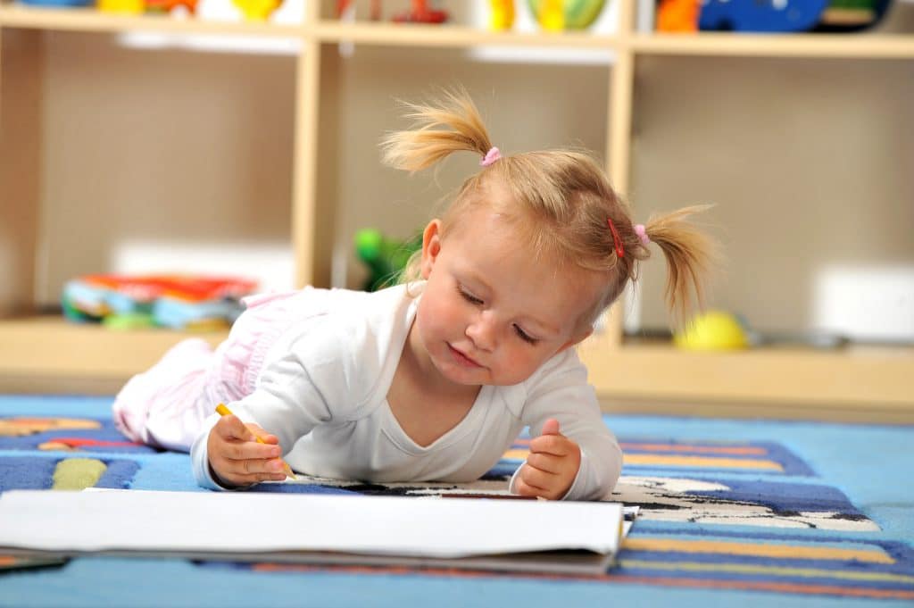 Kleines Mädchen, das auf dem Teppichboden ihres Kinderzimmers liegt und etwas zeichnet.
