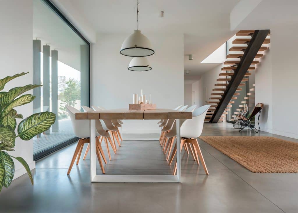 Moderner Essraum mit großem Esstisch aus Holz und acht weißen Lederstühlen neben großflächiger Verglasung und Blick in den Wohnraum.