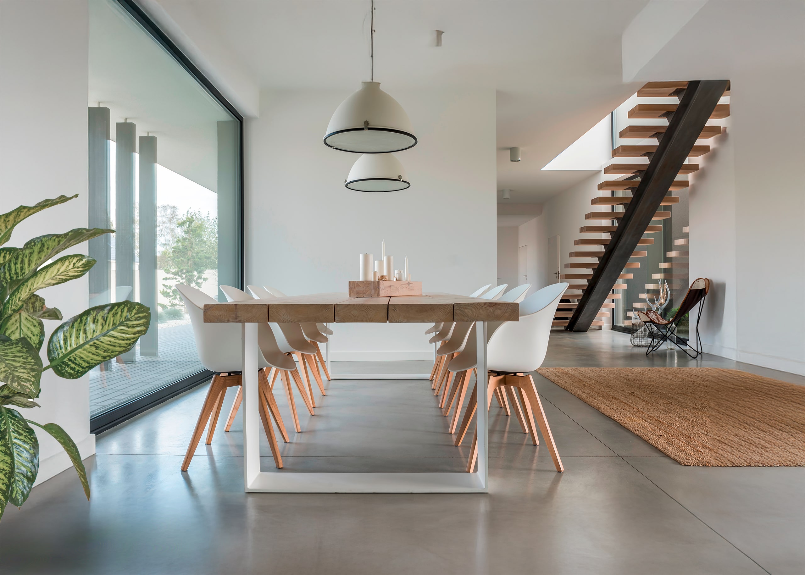 Moderner Essraum mit großem Esstisch aus Holz und acht weißen Lederstühlen neben großflächiger Verglasung und Blick in den Wohnraum.