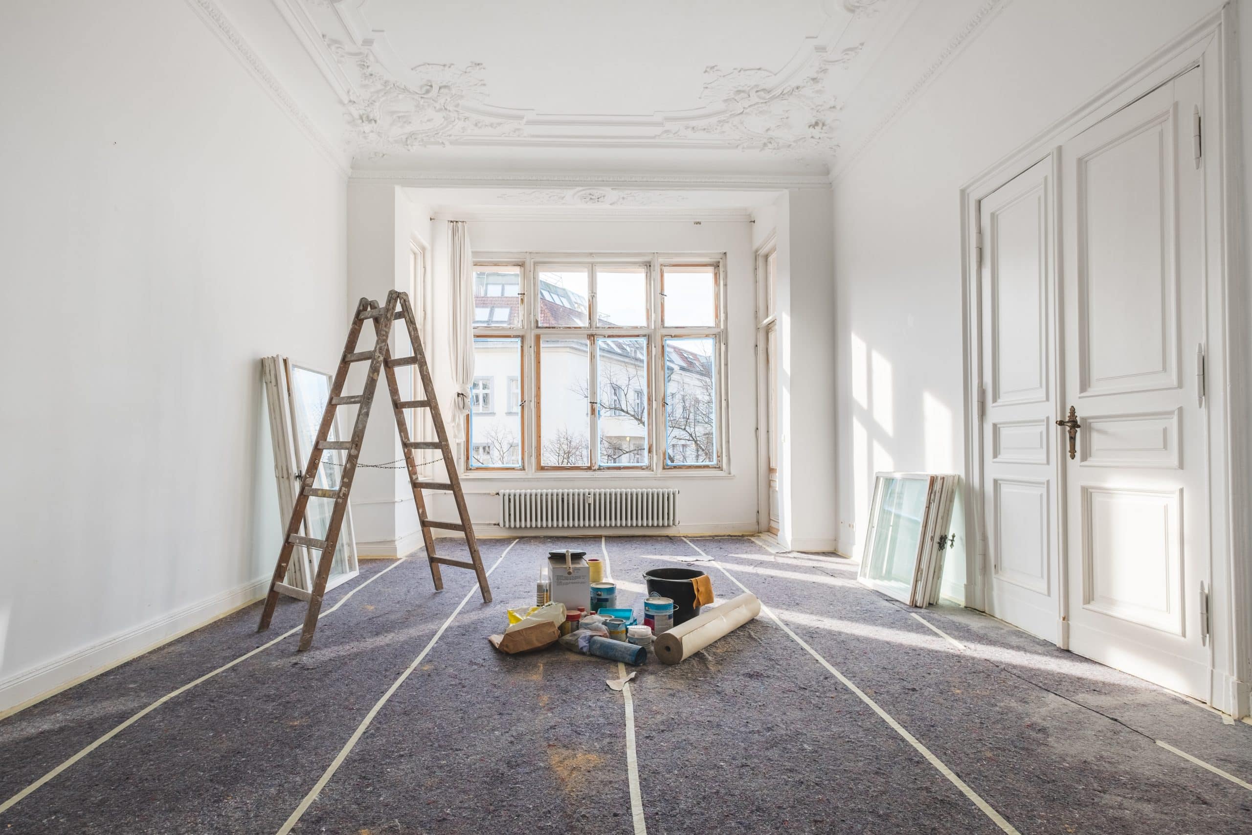 Altbauwohnung mit großen Fenstern, die gerade renoviert und saniert wird. A-Leiter und Utensilien stehen im Raum, der Boden ist mimt Malervlies abgedeckt.