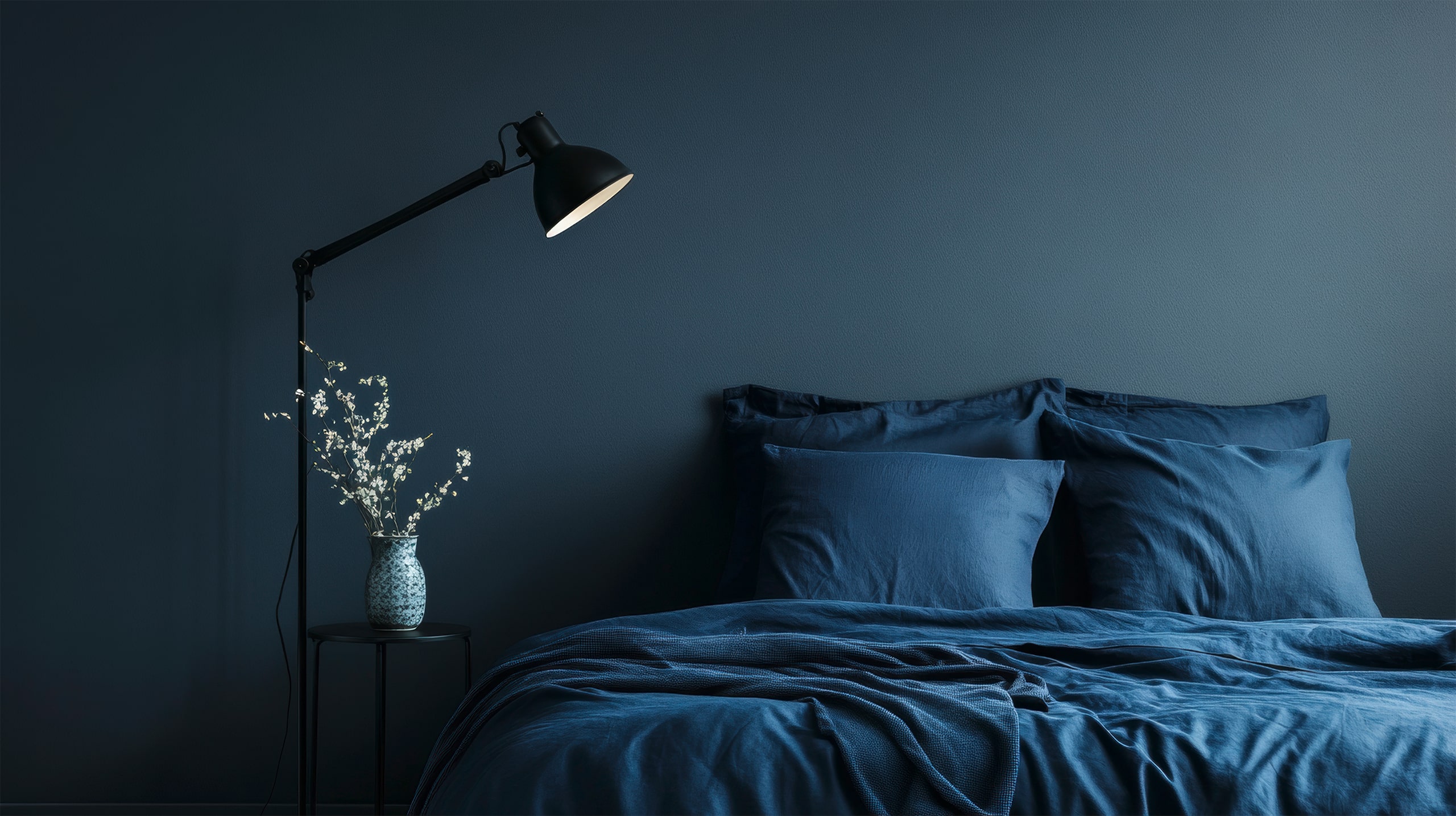 Elegenates Schlafzimmer mit dunkelblauer Wandfarbe und Satin-Bettwäsche in dazu passendem Royalblau. ein keiner, schwarzer Nachttisch mit einer schwarzen Tischleuchte ergänzen den edlen Look.