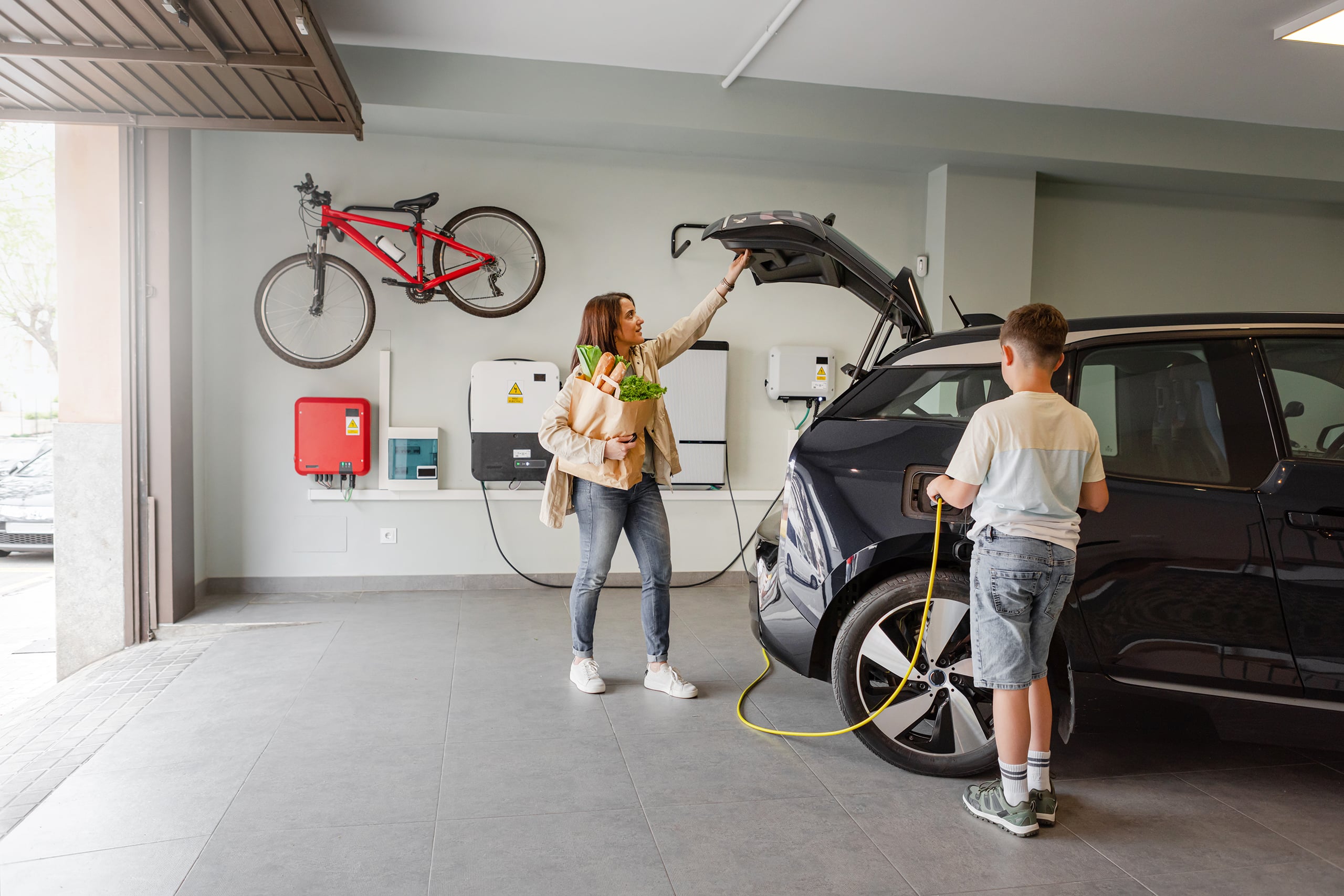 Sohn steckt Elektroauto-Ladegerät von Ladestation an Elektroauto zu Hause, während Mutter tagsüber Lebensmittel aus dem Auto holt. Nachhaltiger alternativer Lebensstil. Horizontaler Kopierraum.