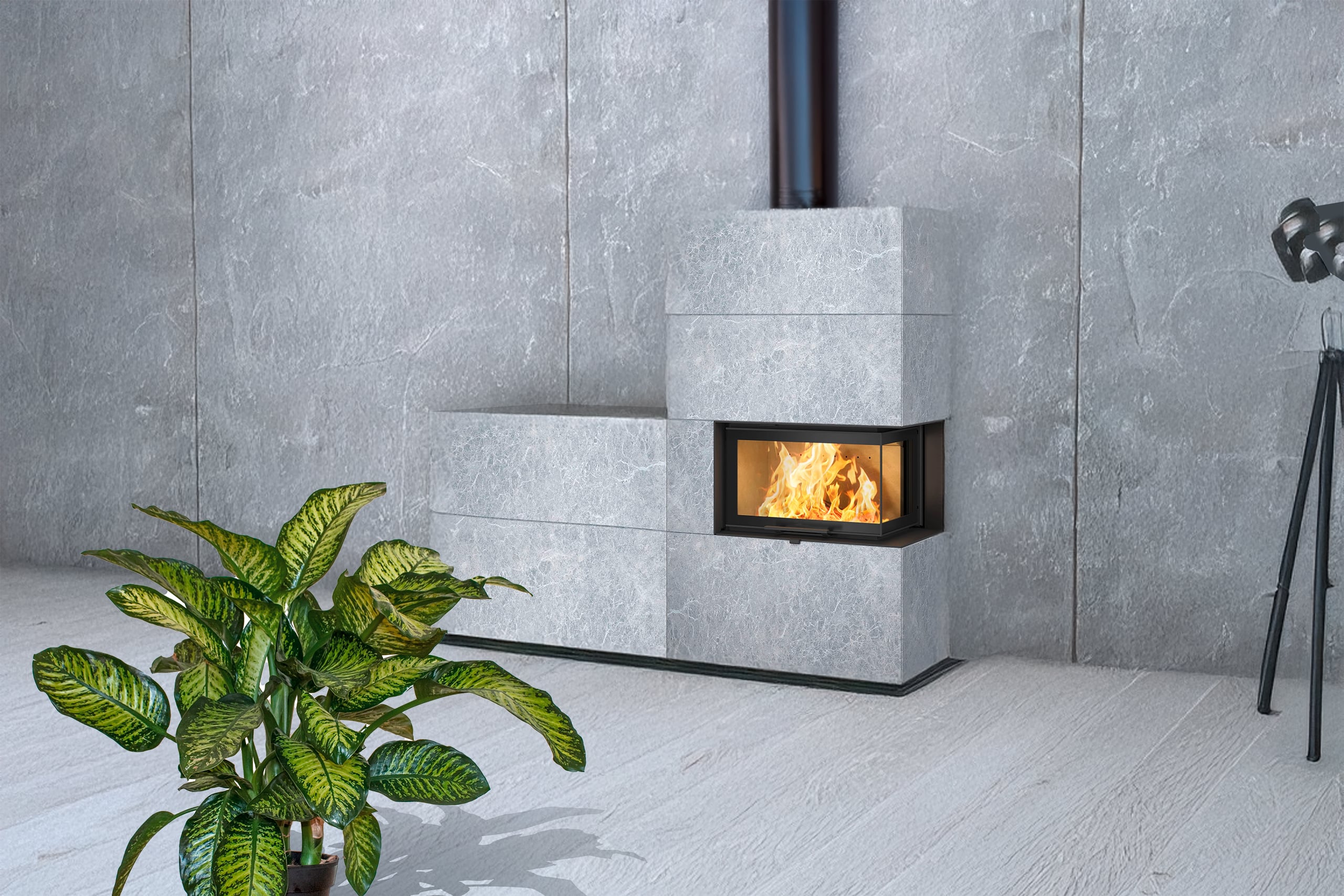 Moderner, freistehender Specksteinofen mit großen, massiven Specksteinplatten, einem Sichtfenster mit brennendem Feuer und einem schwarzen Ofenrohr zur Decke hin, in einer minimalistischen Raumsituation mit Betonrückwand und Grünpflanze im Vordergrund.