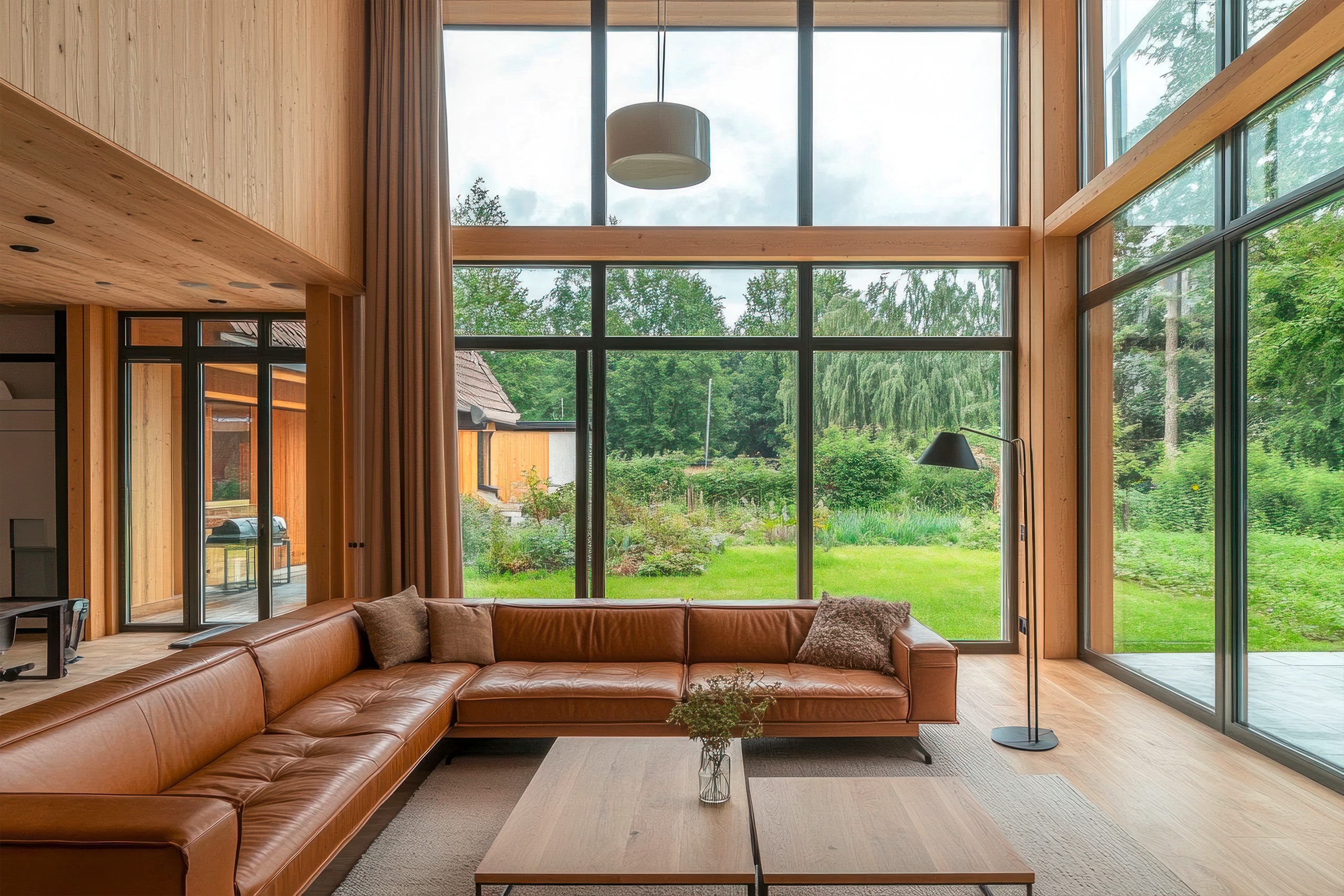 Modernes Wohnzimmer mit großen Fenstern mit Blick in den großzügigen Garten, cognacfärbiges Ledersofa und hellem Holzboden.