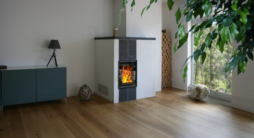 Moderner Kachelofen mit loderndem Feuer durch die Glasscheibe und Holzbriketts zum Nachheizen in einer Wandniesche neben großem Fenste rmit herrlichem Blick in die grüne Natur.