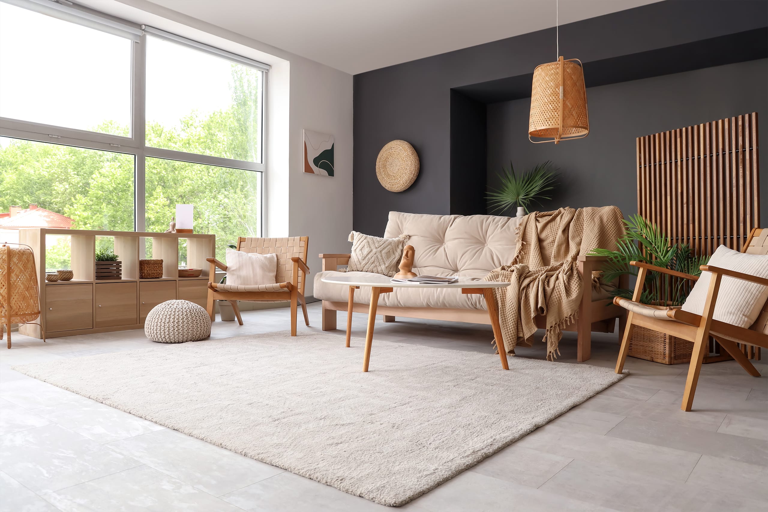 Gemütliches Sofa aus Holz mit bequemen Sitzpolsern in beige, dazupassenden Sesseln und Nieren-Couchtisch im Wohnzimmer. Ein freiliegender Teppich in creme, ansprechende Accessoires und eine Rückwand in Anthrazit runden den einladenden Wohnraum ab.