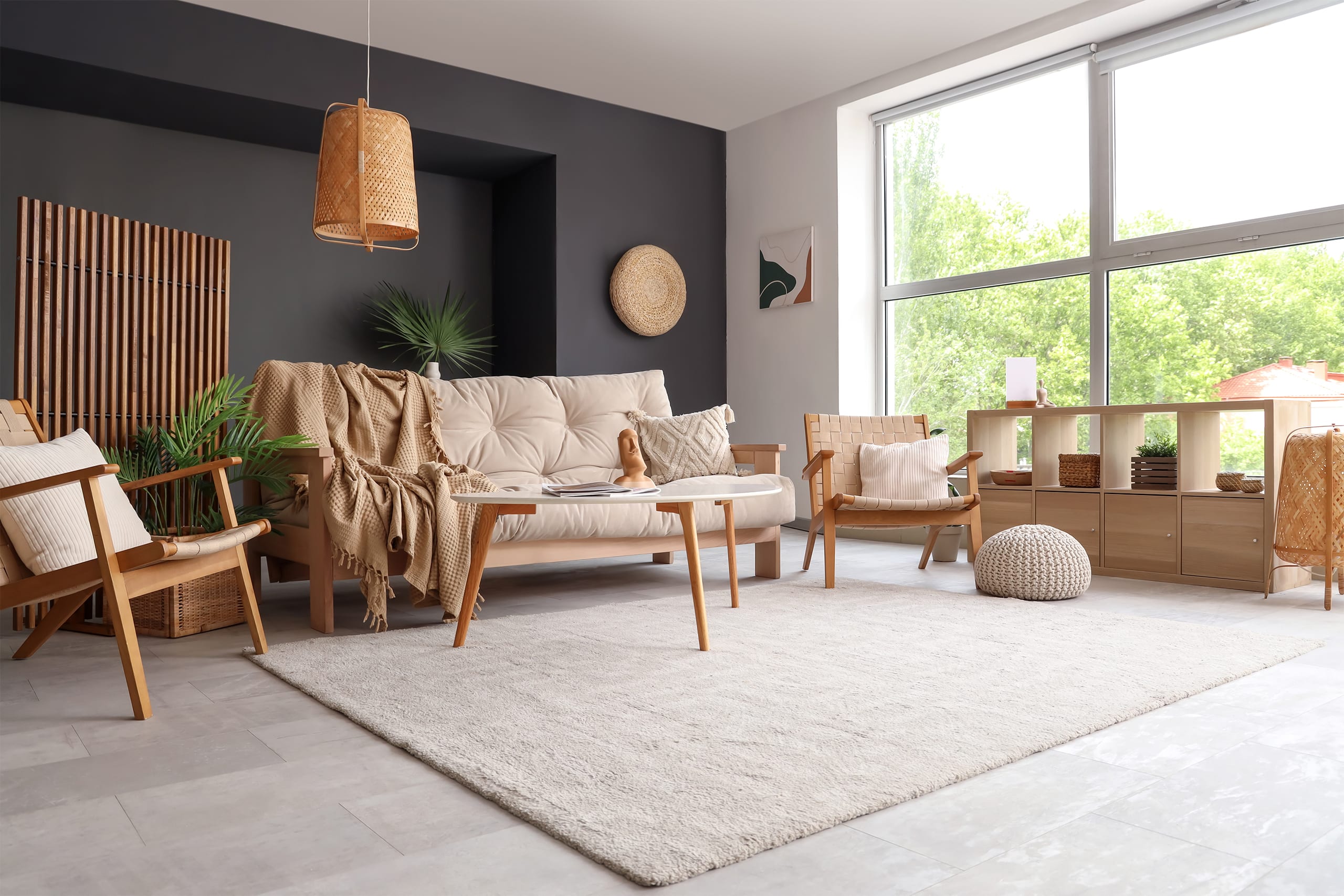 Gemütliches Sofa aus Holz mit bequemen Sitzpolsern in beige, dazupassenden Sesseln und Nieren-Couchtisch im Wohnzimmer. Ein freiliegender Teppich in creme, ansprechende Accessoires und eine Rückwand in Anthrazit runden den einladenden Wohnraum ab.