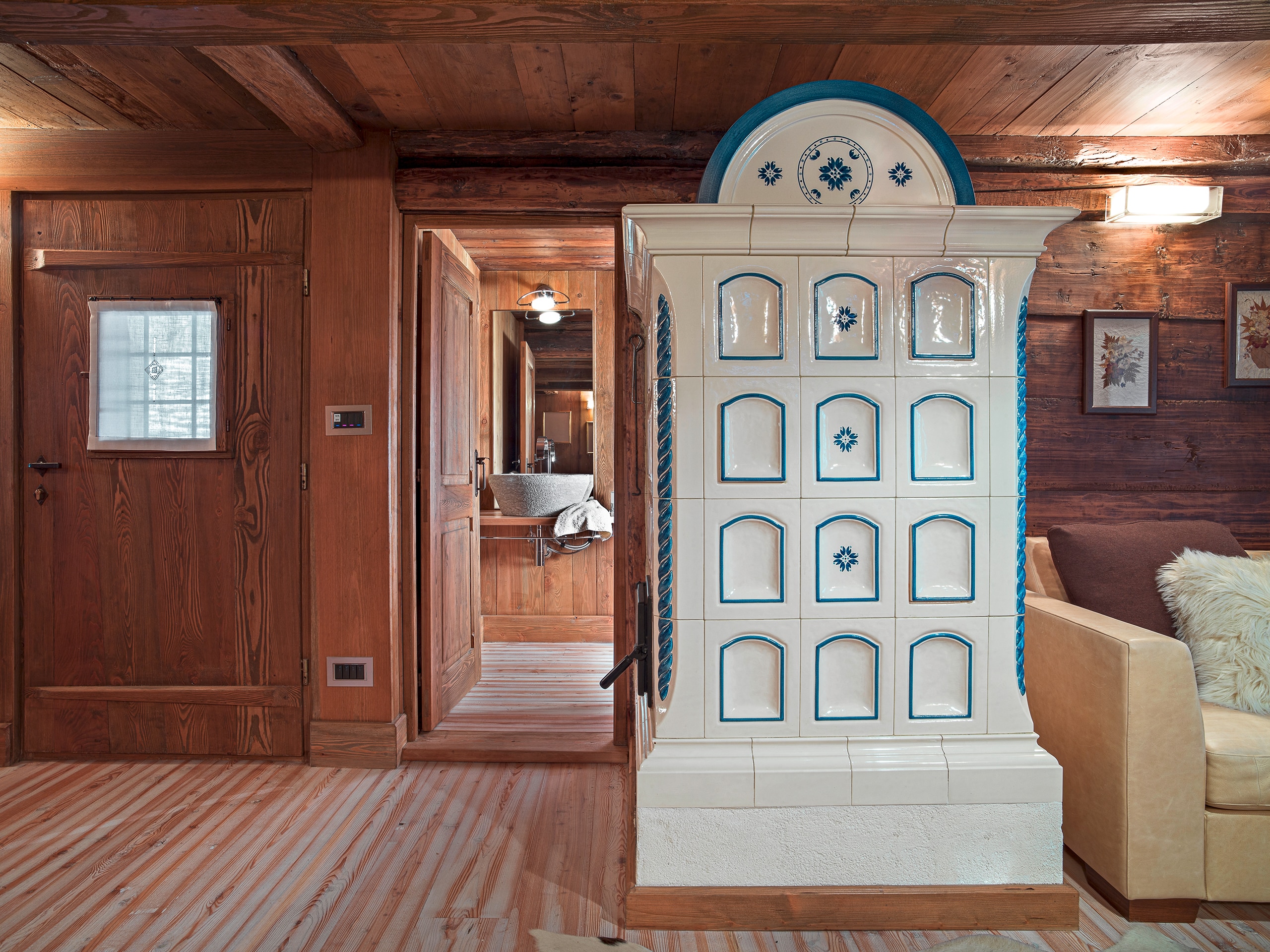 Innenaufnahme eines rustikalen Wohnzimmers - im Vordergrund der typische Kachelofen in weißer und blauer Glasur und aufwendigem Design neben der Eingangstür. Boden, Decke und Wände sind aus Altholz.