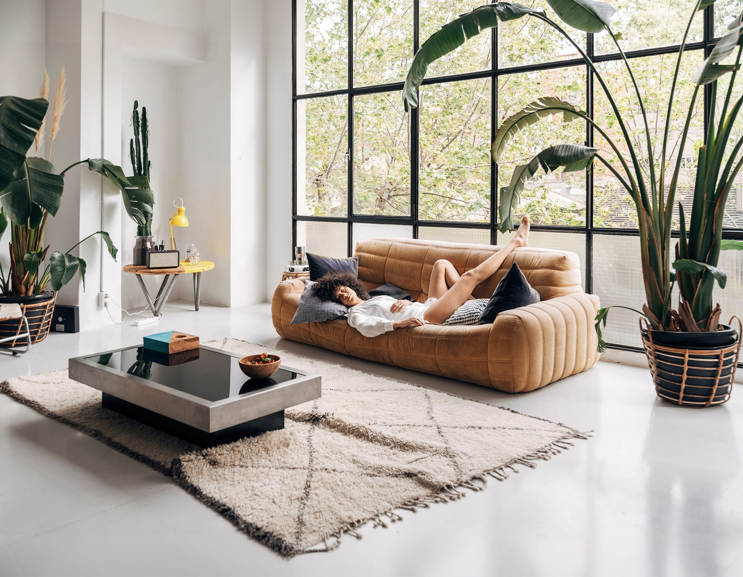 Junge Frau auf einem cognacfarbenen Ledersofa, einem Wohnzimmertisch samt naturfatbenen Teppich darunter in einem großen, hellen Wohnbereich eines Lofts mit großen Pflanzen und minimalistischen Mobilar schläft gemütlich.