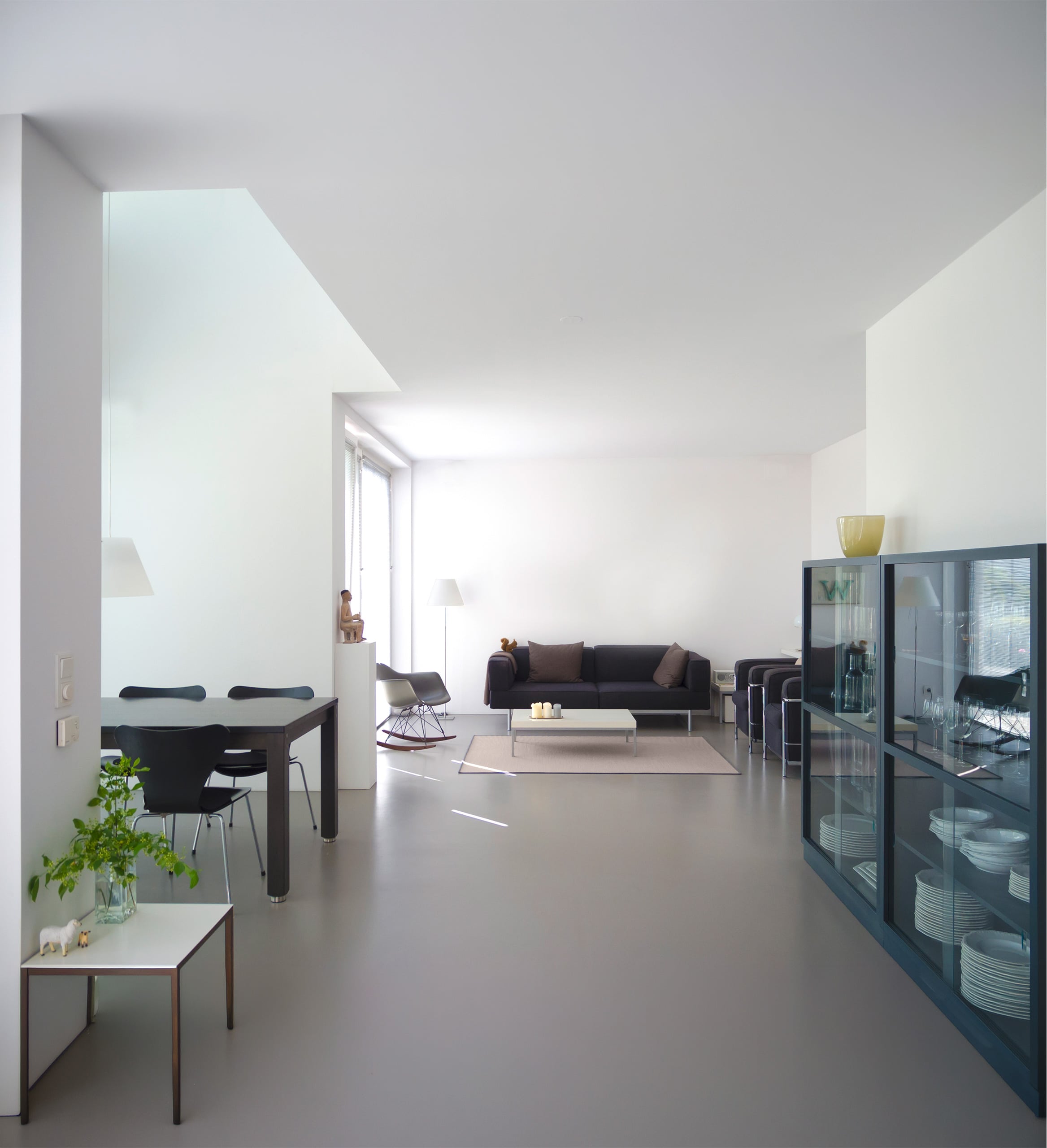 Modernes, schlichtes Wohnzimmer mit weißen Wänden, grauem Sichtestrich (Designestrich) und akzentreichen Accessoires, wie eine gelbe Vase und eine Holzfigur.