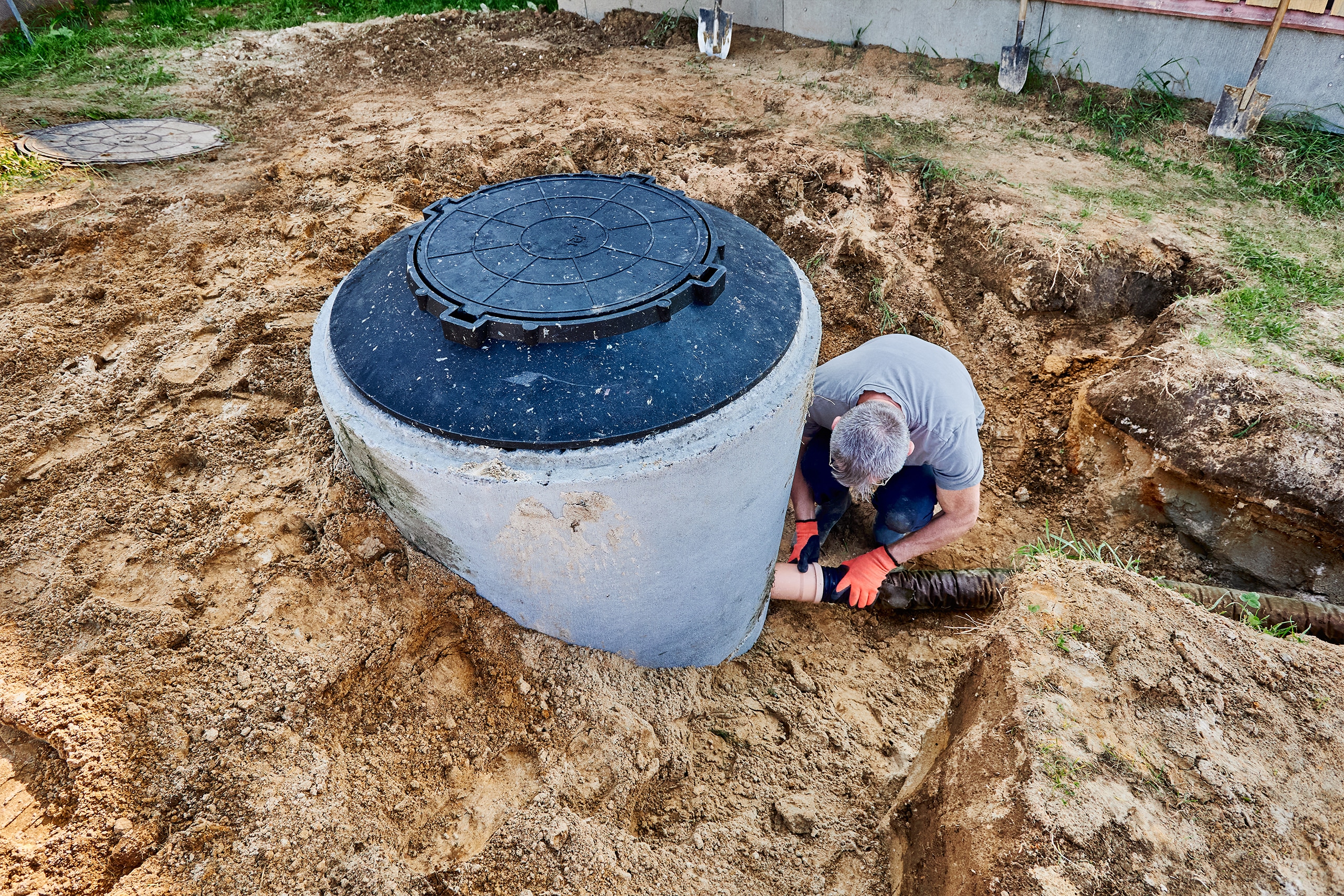 Ein Klempner (Installateur) schließt das Abwasserrohr an das Auslassrohr der Klärgrube (Senkrgrube) an, um das Abwasser zum Versickerungsfeld zu leiten.