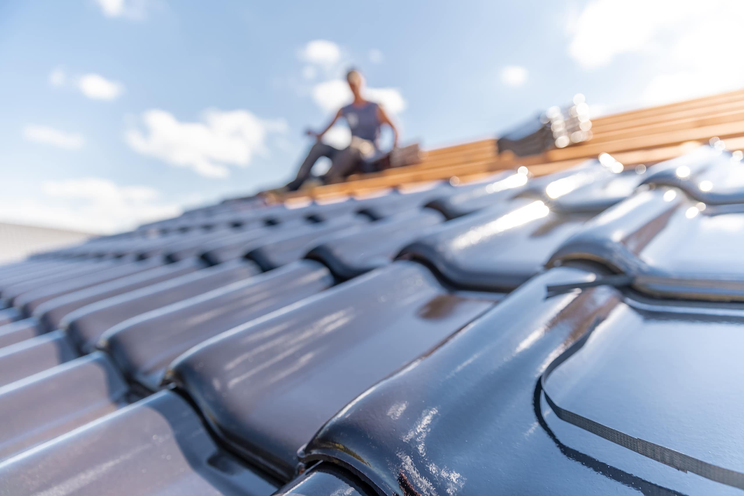 Dachdecker mitten im Decken eines Daches mit gebrannten, schwarzen Keramikziegel auf dem Dach. Die bisher verlegten Ziegeln sind dabei im Vordergrund.