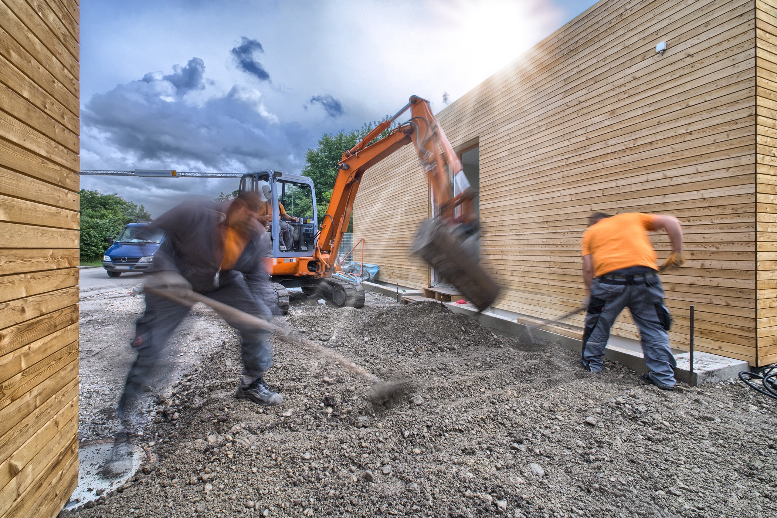 Arbeiter sind auf der Baustelle eines modernen Neubauhauses mit Flachdach und Holzfassade mit Erdarbeiten bzw. Gartengestaltung / Landschaftsbau beschäftig. Ein Bagger im Hintergrund gräbt dabei Schotter und Erde in Richtung der Arbeiter.