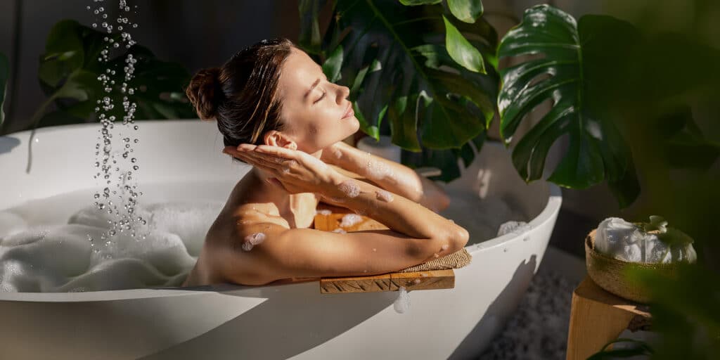 Junge Frau genießt eine entspannte Zeit in der Badewanne mit warmen Wasser und Badeschaum in Ihrem Badezimmer. Umgeben von sanftem, natürlichem Licht und einer entspannenden Atmosphäre mit tropischem Grün. - KI-generiert