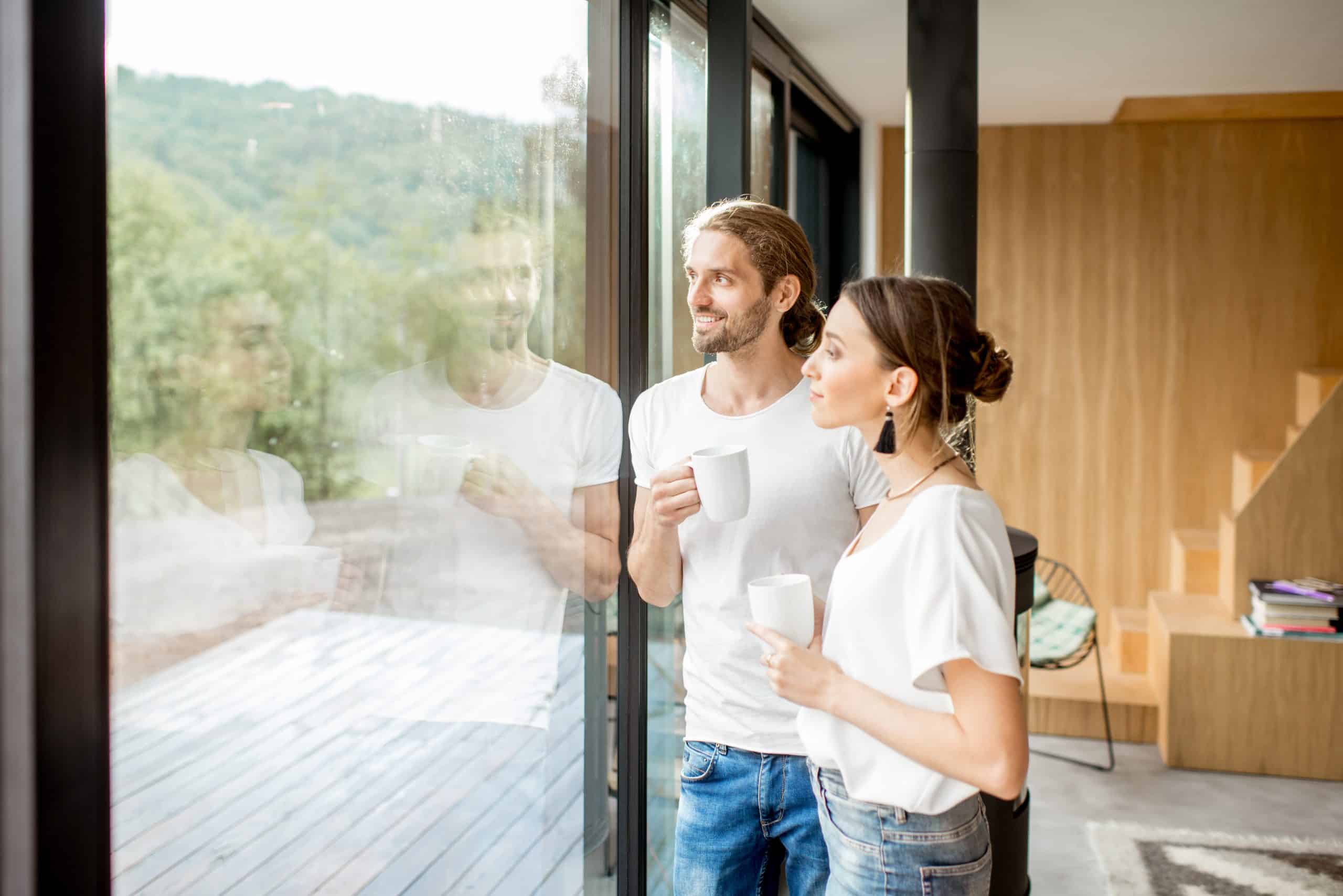 Junges paar genießt eine Tasse Kaffe mit Blick aus dem modernen Schiebefenster des neuen, modernen Zuhauses.