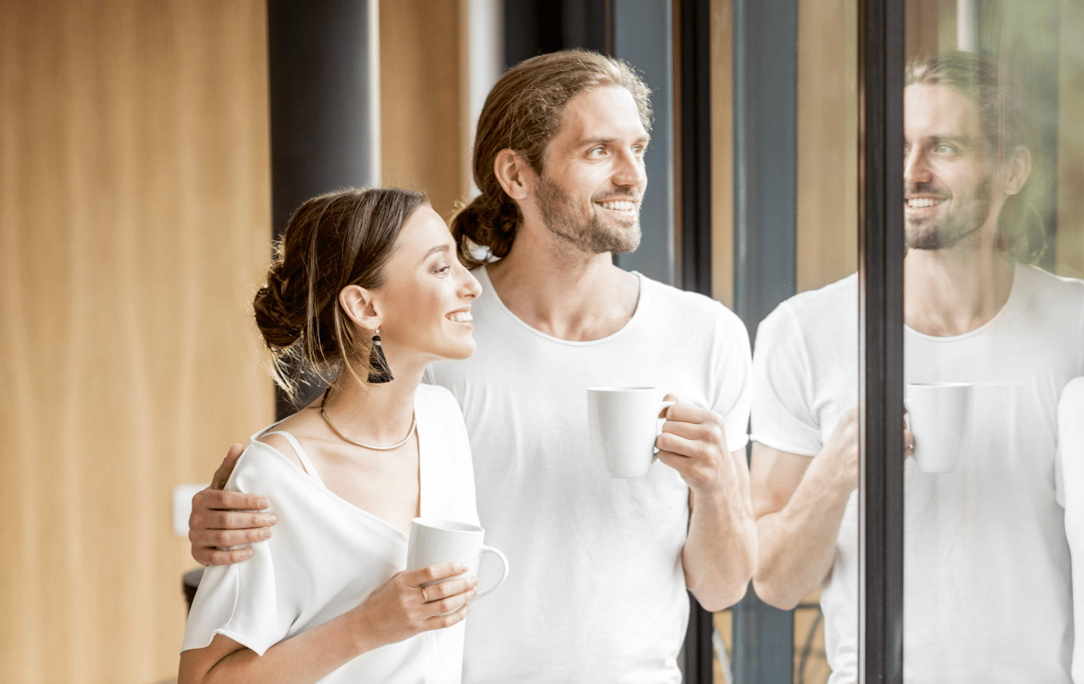 Junges Paar steht mit Kaffee am Fensters Ihres modernen Hauses und blicken nach Draußen.