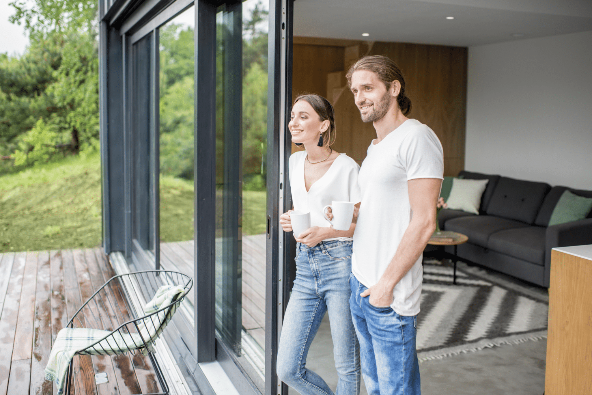 Junges Paar genießt bei einer Tasse Kaffee den Ausblick auf die Terrasse mit Garten Ihres neuen, modernen Hauses mit großen Schiebetüren in Anthrazit.