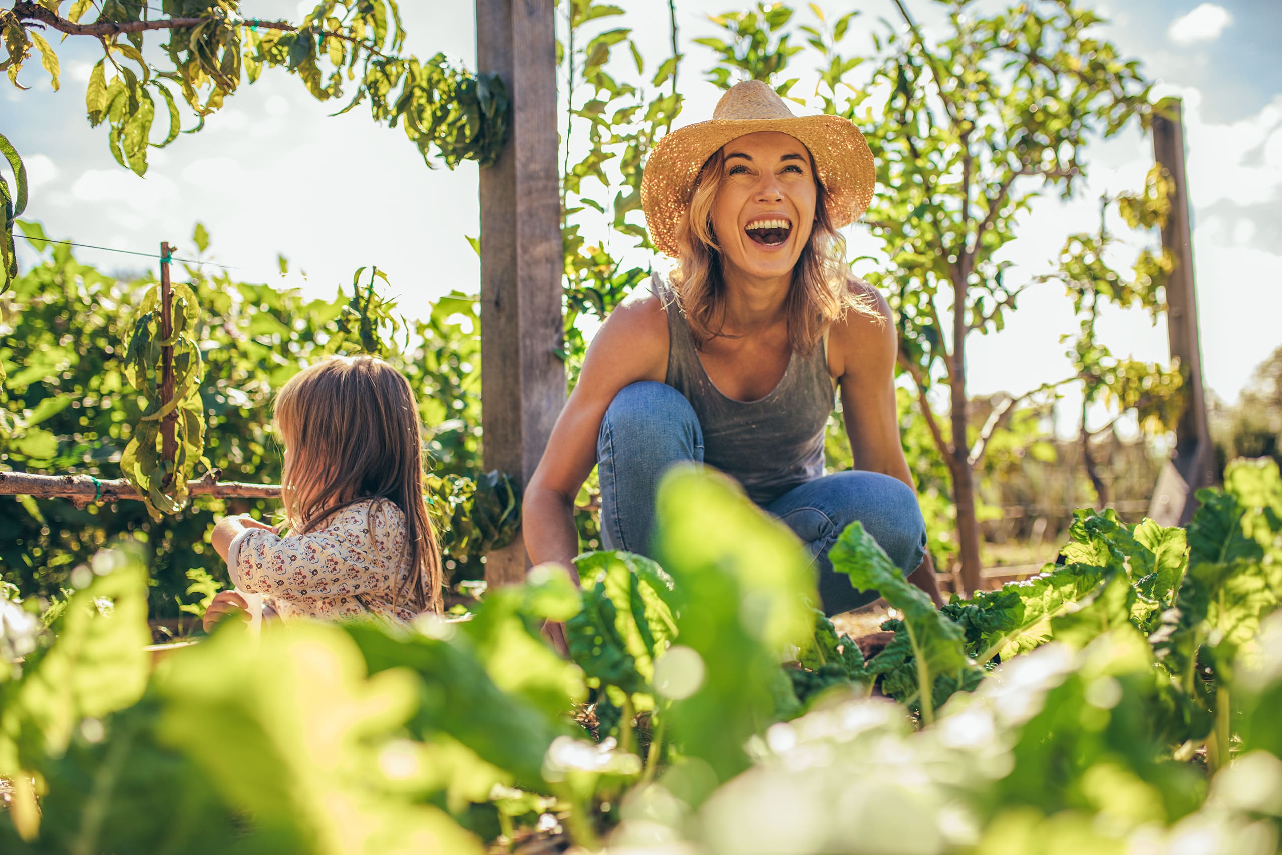 Glücklich lachende Frau mit Ihrer kleinen Tochter kniet im Gemüsegarten und freut sich über die üppige Ernte und den sonnigen Tag.