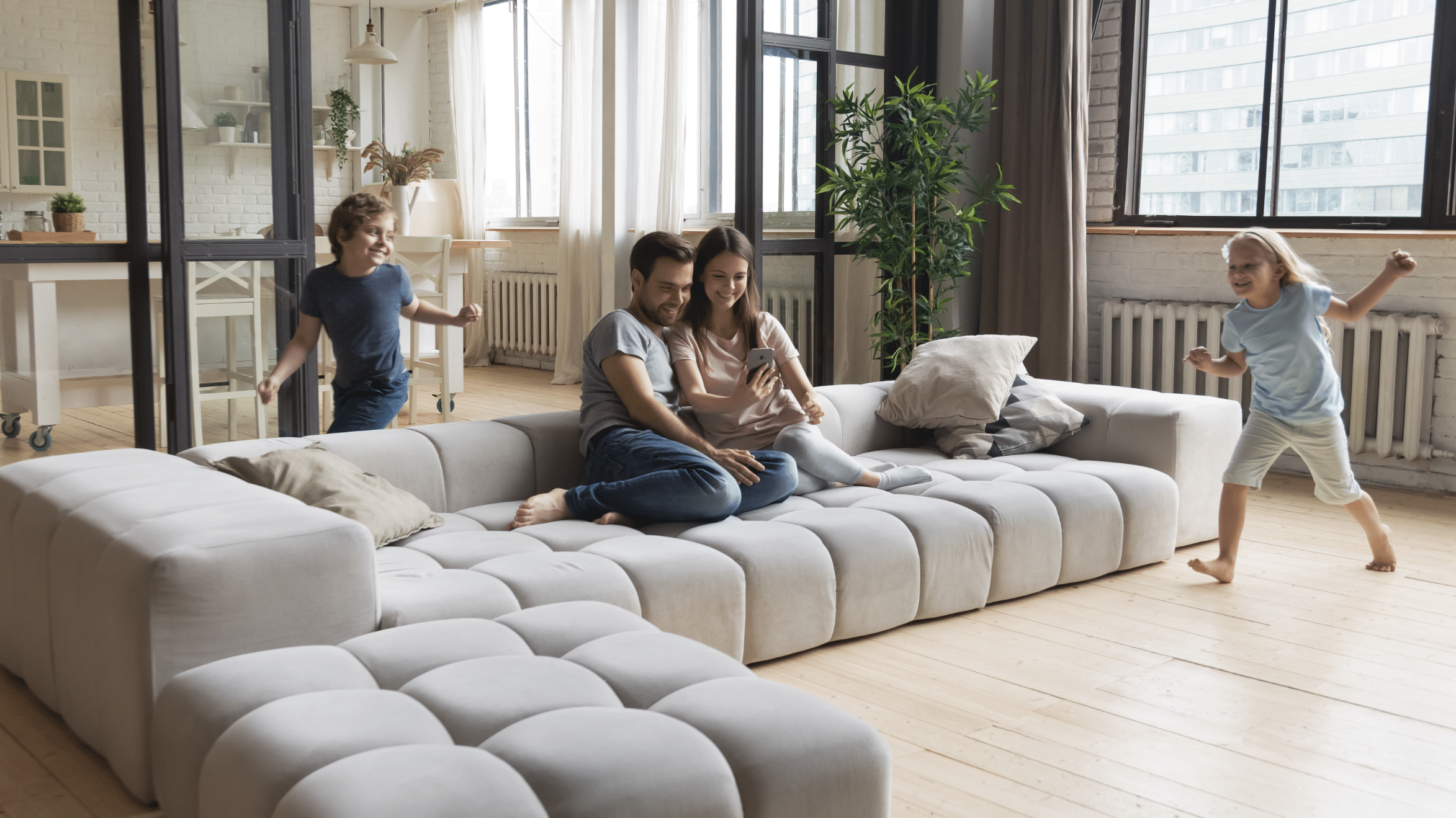 Junge Familie genießt die gemeinsame freie Zeit in der Altbauwohnung mit Loft-Charakter. Die Eltern machen ein Selfie am Mobiltelefon, während die Kinder um das große, hellgraue Wohnzimmer-Sofa laufen.