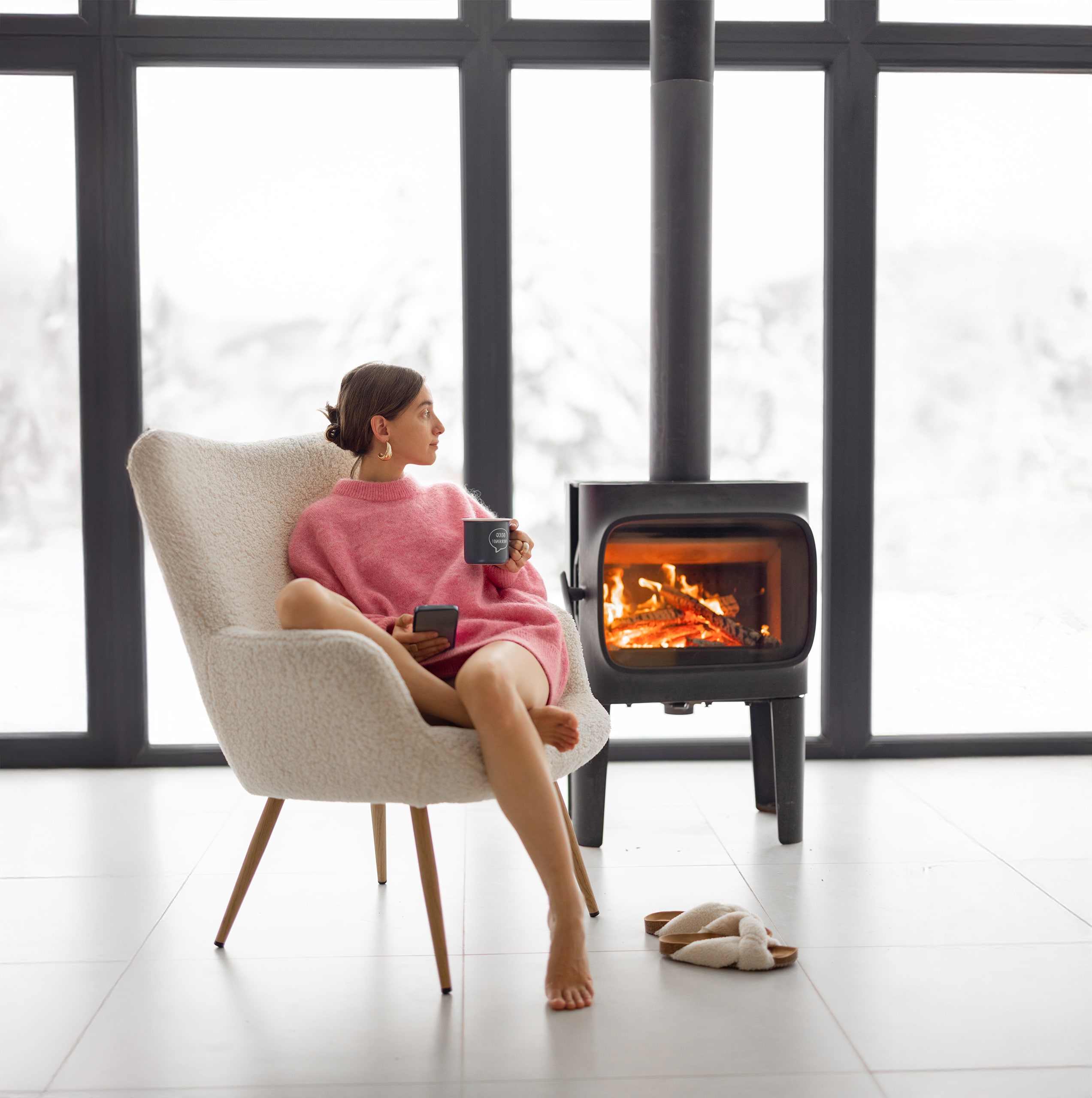 Junge Frau sitzt mit einer Tasse Tee auf einem gemütlichen Loungesessel am freistehenden Kamin mit Sichtfenster zum lodernden Feuer in einem modernen Haus inmitten der Natur.
