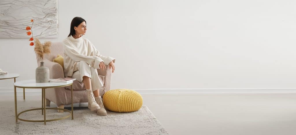 Junge Frau sitzt in einem beigen Sessel und elegenaten Accessoires, neben einer hellen und freien Wand.