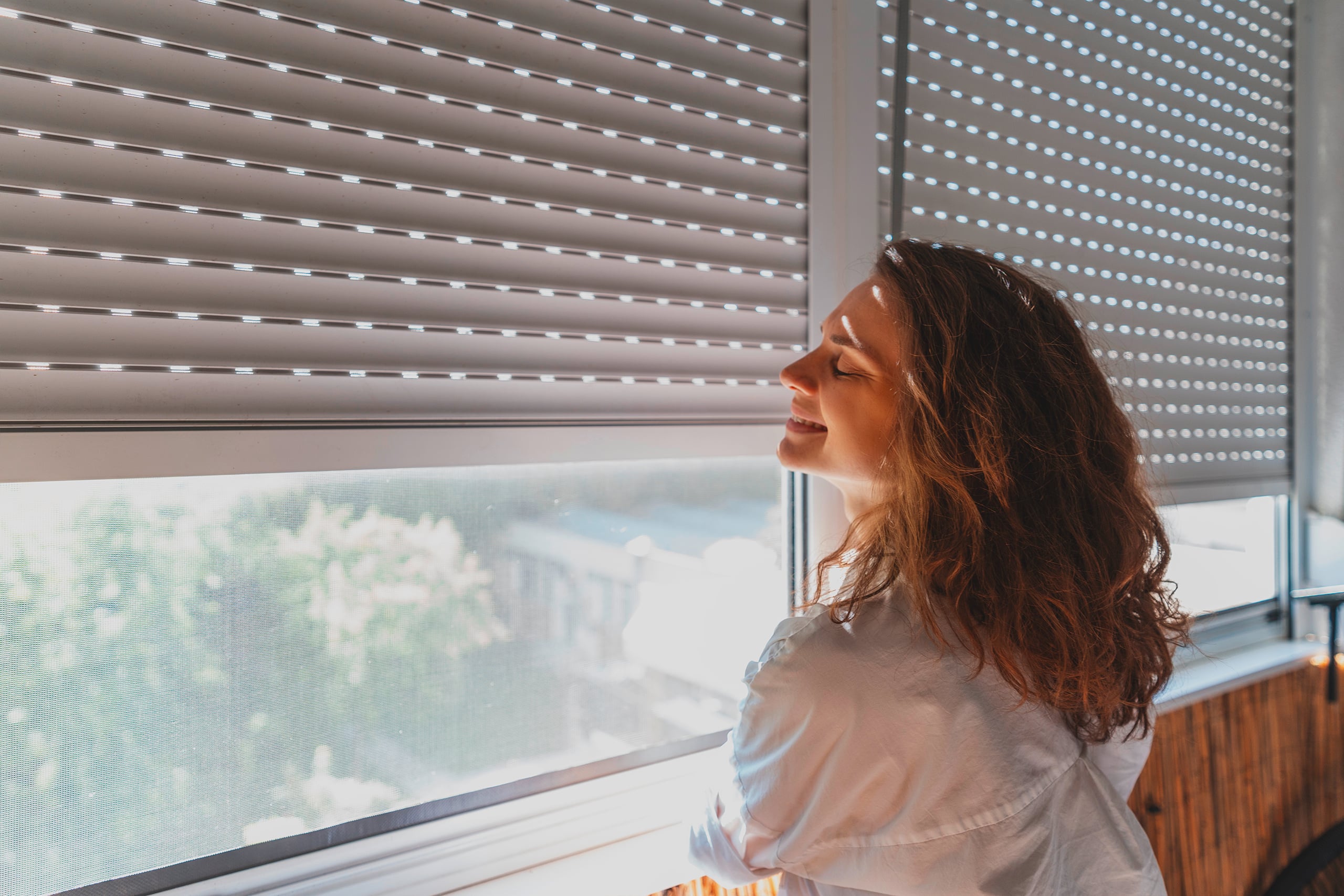 Junge Frau öffnet an einem Sommertag die Rollläden auf dem Balkon.