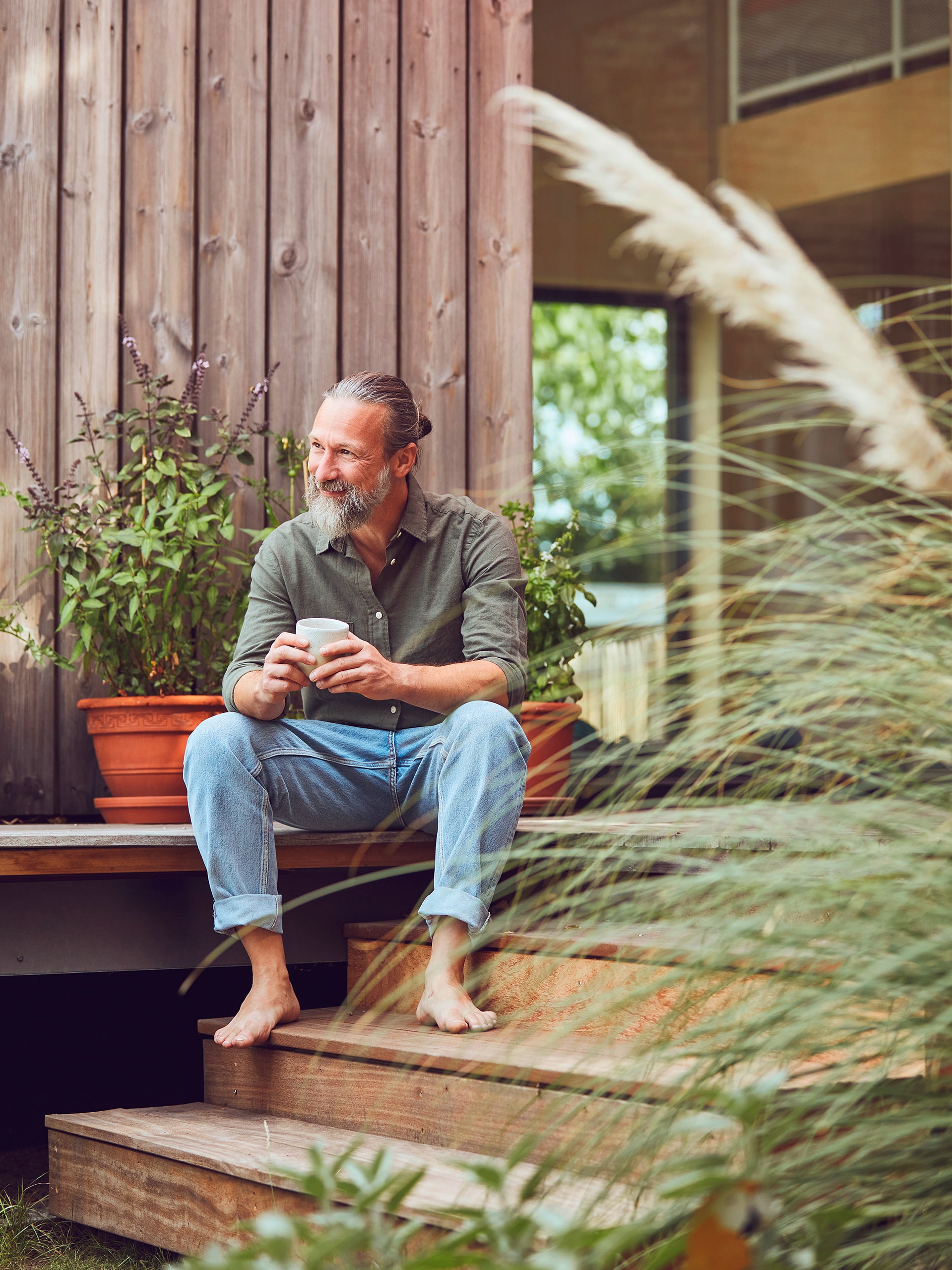 Zufrieden lachender, reifer Mann im Hipster-Look hält Kaffeetasse, während er auf den Stufen seines Tiny Houses sitzt.