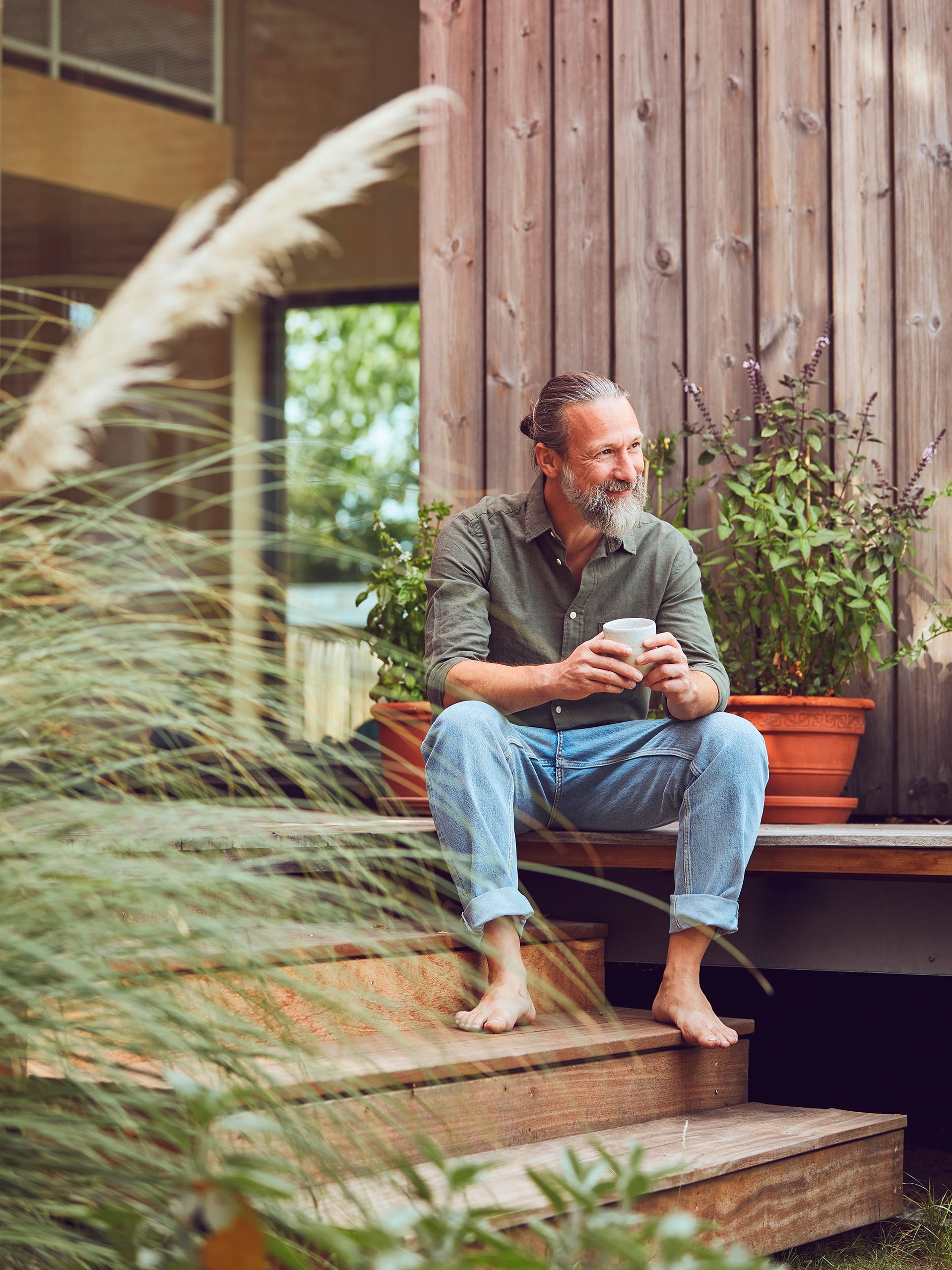 Zufrieden lachender, reifer Mann im Hipster-Look hält Kaffeetasse, während er auf den Stufen seines Tiny Houses sitzt.