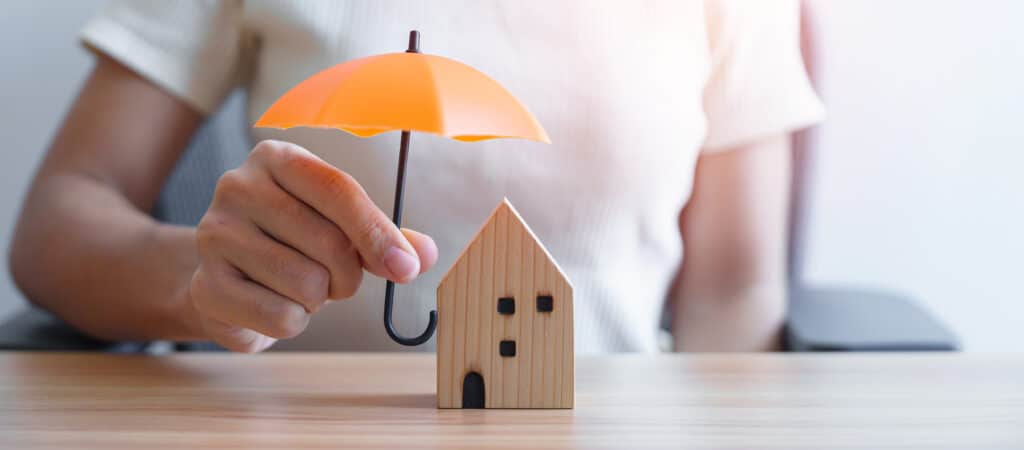 Hand einer Geschäftsfrau hält einen orangefarbenen Regenschirm über ein hölzernes Hausmodell – Konzepte aus den Bereichen Immobilien, Versicherungen und Eigentum.