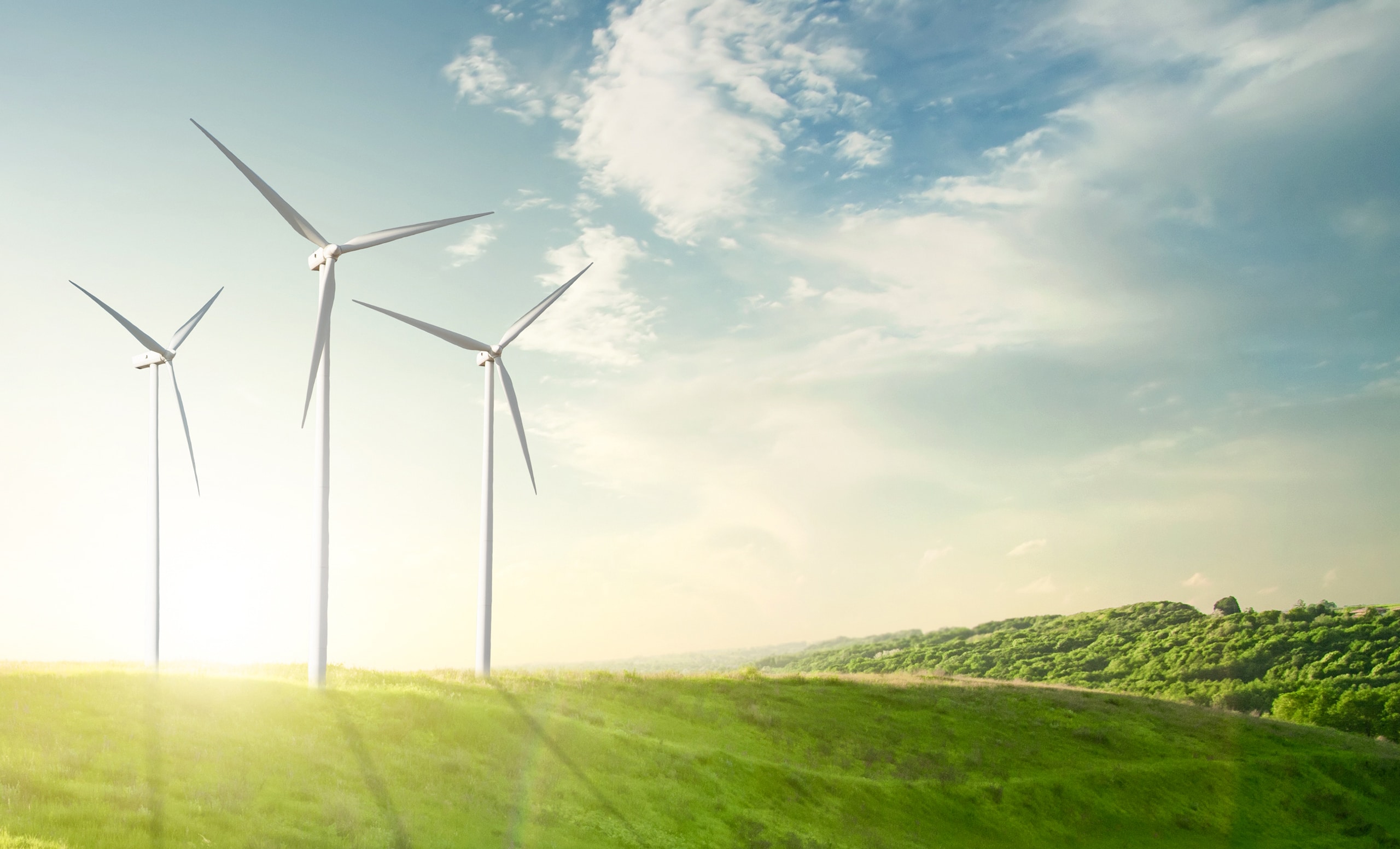 3 Windräder auf der Anhöhe eines Hügels einer sanft hügeligen, unbewohnten Landschaft.