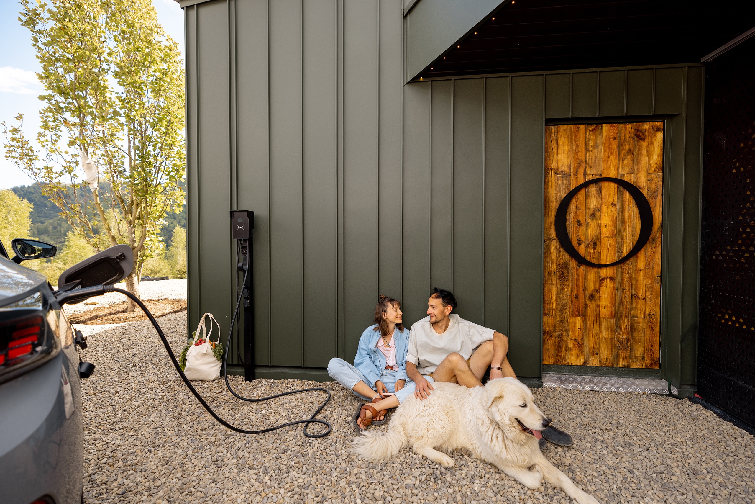 Junges Paar sitzt mit seinem Hund in der Nähe eines modernen Hauses, in der Nähe lädt ein Elektroauto. Konzept der Liebe, des modernen Lebens und des Wohlbefindens.