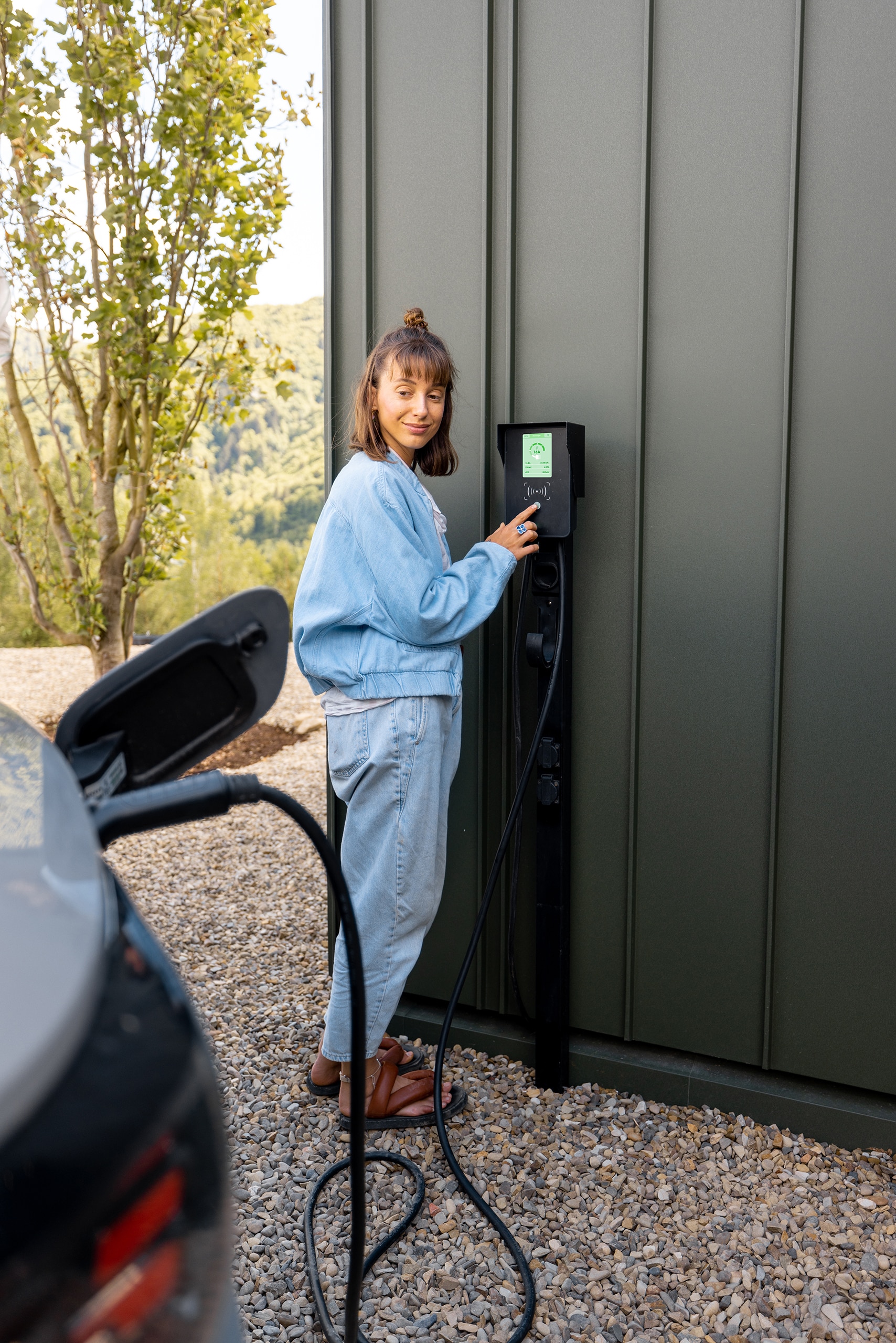 Frau schaltet Autoladestation ein, während sie ihr Elektrofahrzeug direkt mittels Wallbox an der Hausfassade auflädt und nutzt dieses innovative Konzept intelligenter Technologien für komfortables Leben mit Elektroautos.